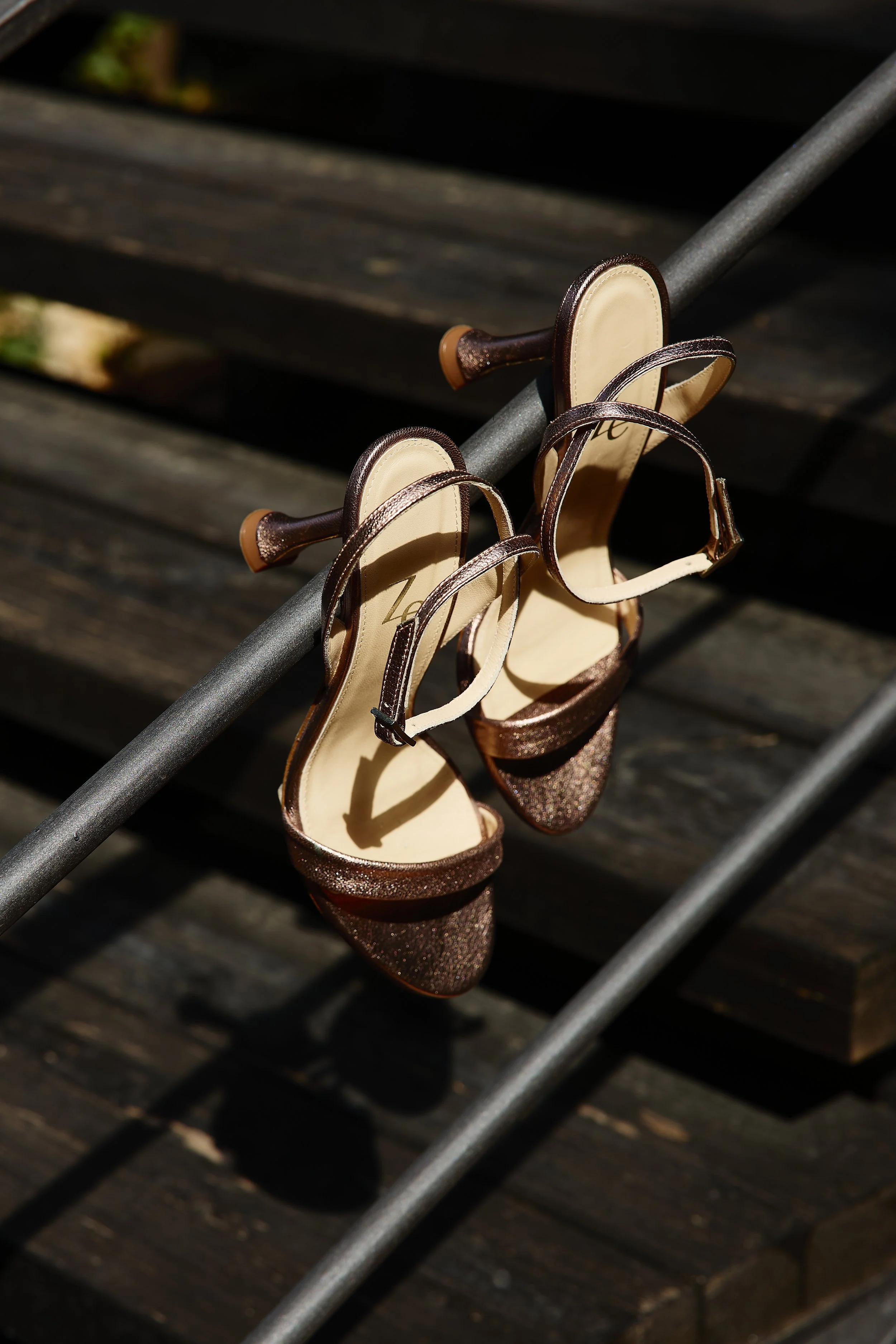 Zapatos de tacón con grabados dorados colgados en una baranda de metal sobre un banco de madera.