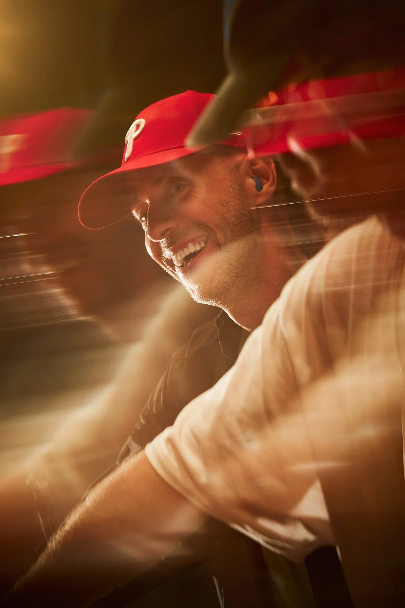 Hombre sonriendo con gorra roja y auriculares azules, en un ambiente con luces cálidas.
