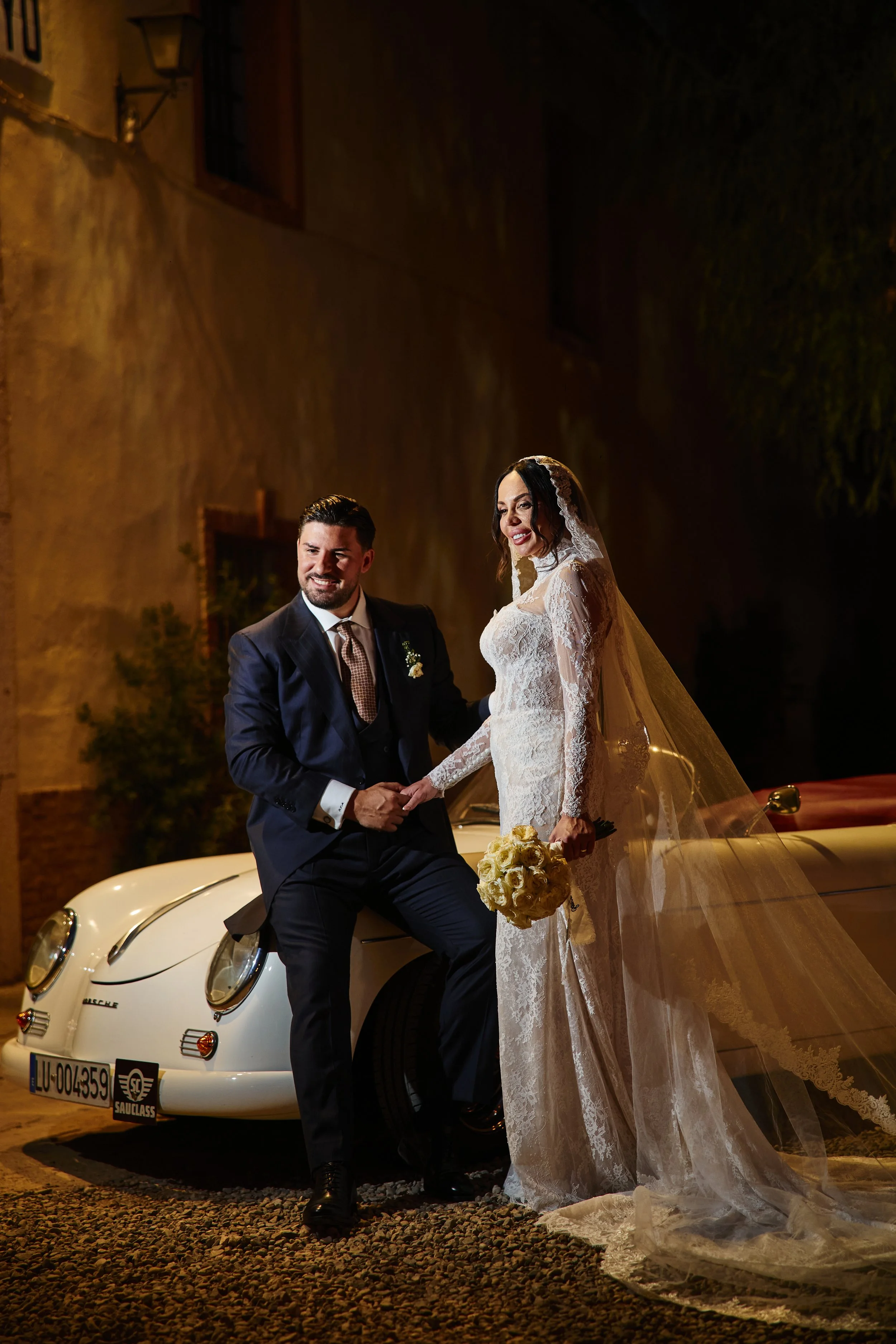 Pareja de recién casados posando con una casa antigua y un coche clásico Porsche en la noche. Ella lleva vestido de novia y él traje formal con corbata. Ella sostiene un ramo de rosas blancas.