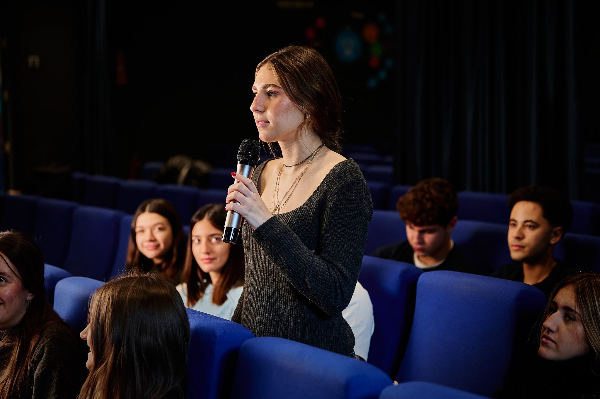 Joven mujer de cabello castaño claro levantando la mano en una audiencia en un cine o teatro oscuro, con varias personas sentadas en las butacas azules alrededor.