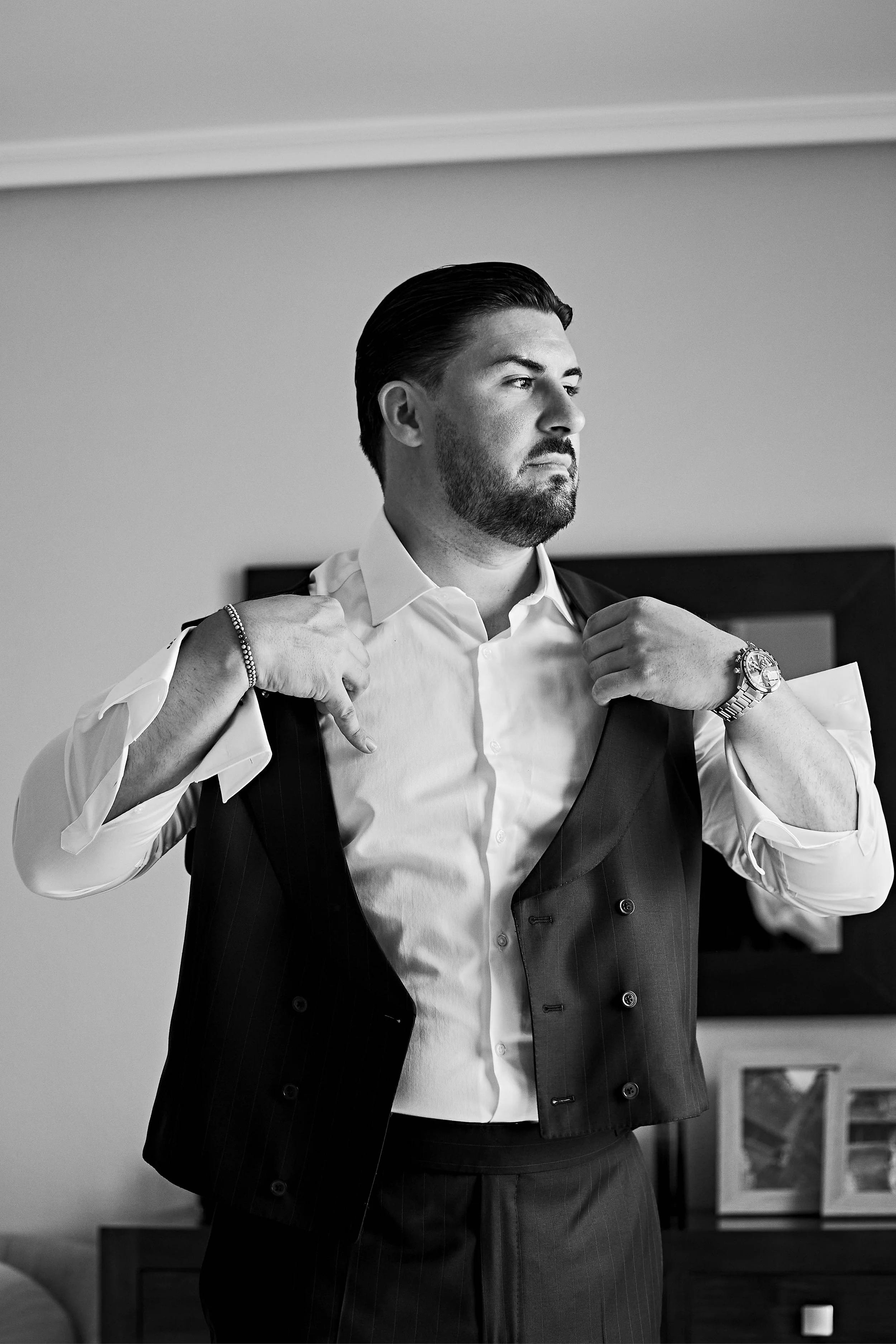 Un hombre con barba arreglada y cabello peinado hacia atrás se está vistiendo con una camisa blanca y un chaleco a rayas, mientras se coloca el saco. Tiene un reloj en la muñeca izquierda y una pulsera en la derecha. traje dee sastrería diplomatica
