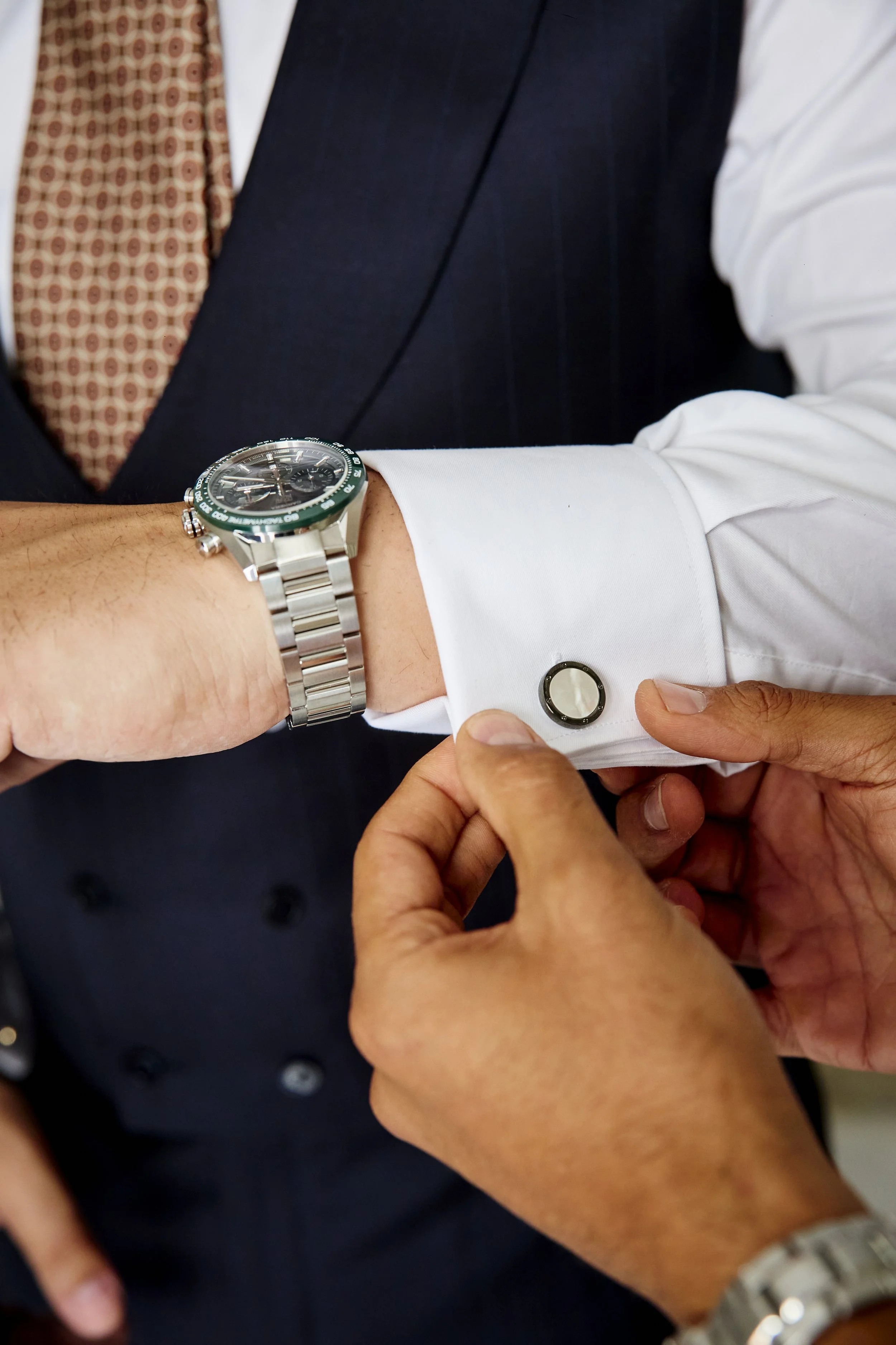Persona colocando un gemelo en la manga de una camisa blanca, con un reloj de pulsera en la muñeca y vestimenta formal, incluyendo un saco y corbata.