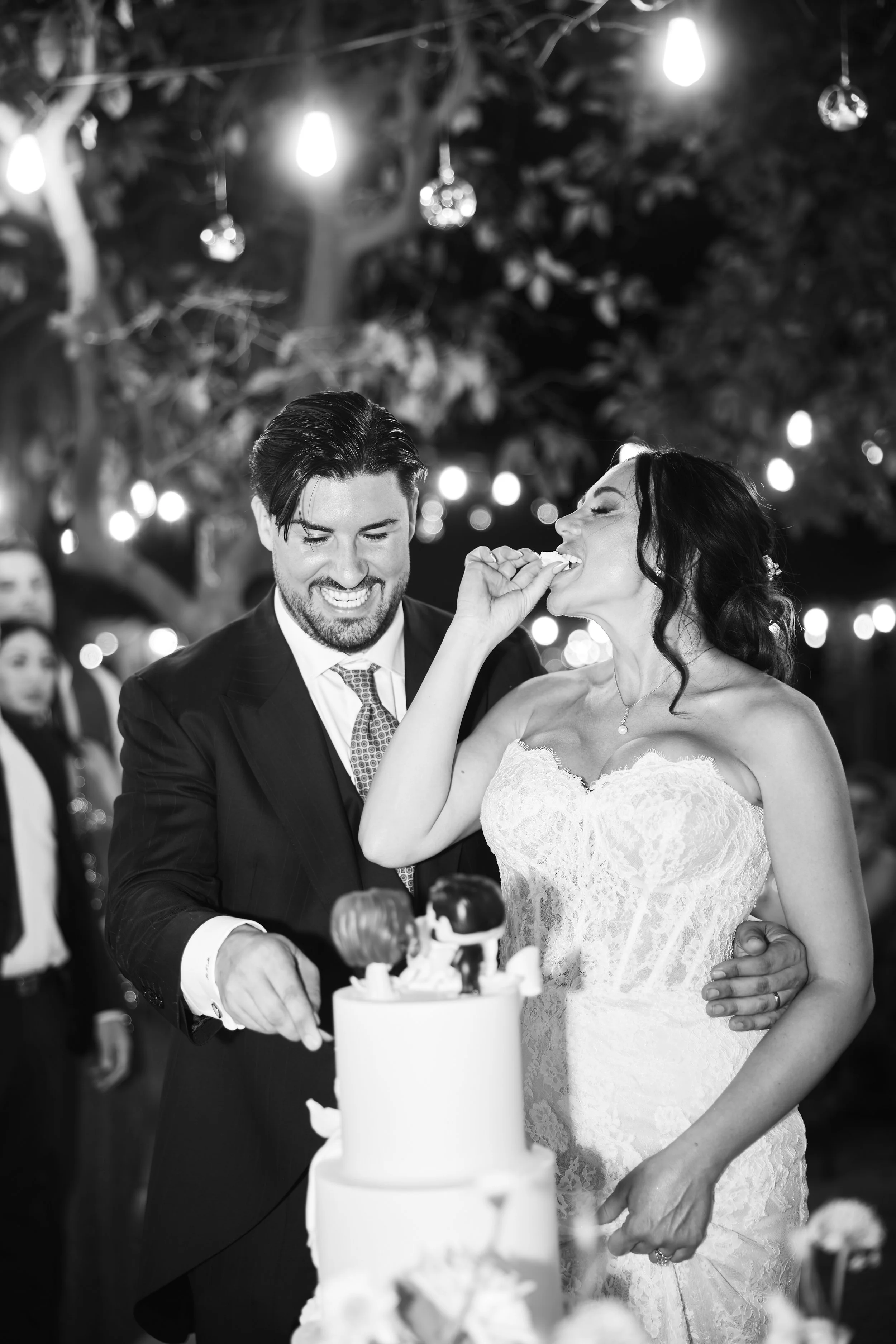Pareja de novios en una boda cortejándose y sonriendo mientras cortan un pastel de bodas en una celebración nocturna con decoraciones y luces en el fondo.