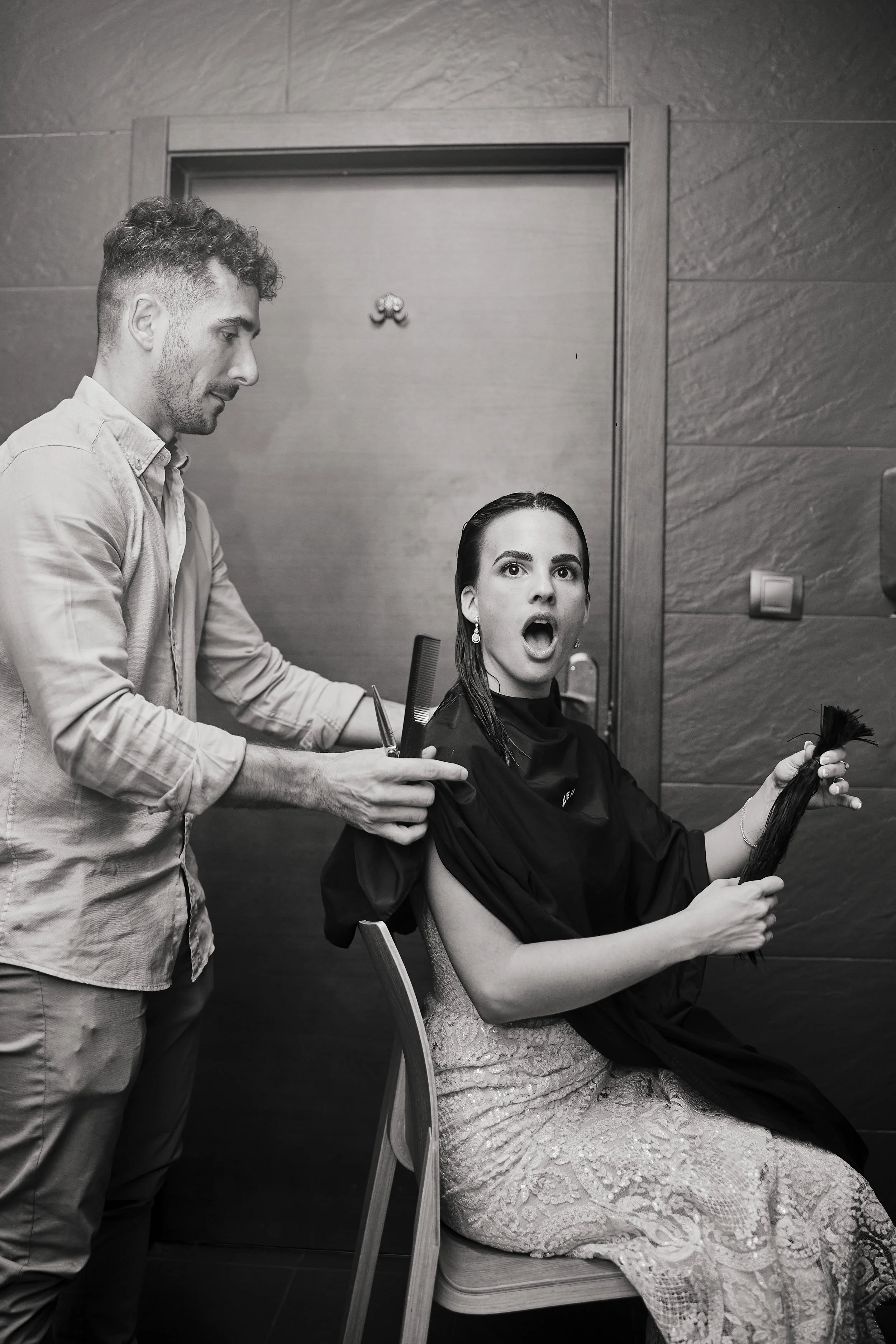 Mujer en una sala de belleza, sentada en una silla, con el cabello mojado y una expresión sorprendida, mientras un estilista le corta el cabello y sostiene un mechón.