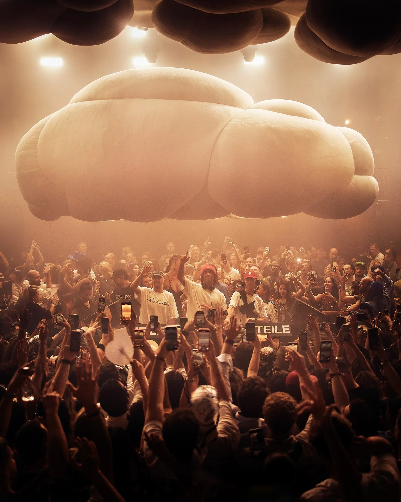 Concierto en interiores con un escenario y una gran multitud de personas, muchas con teléfonos en mano, y una estructura de nubes inflables en el techo.