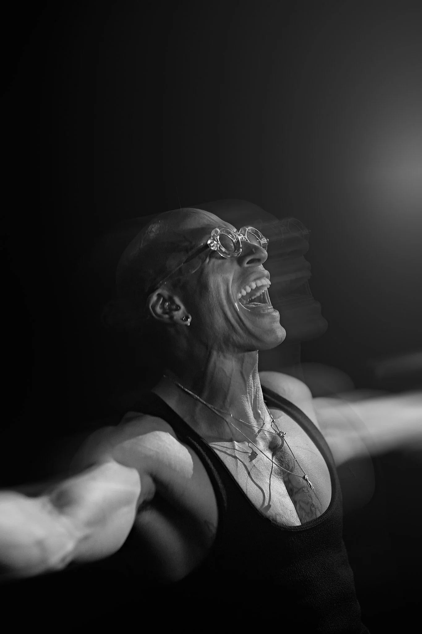 Una mujer con gafas y peinado corto, en un fondo oscuro, se está tomando una selfie con expresión de alegría y risa, en una foto en blanco y negro.