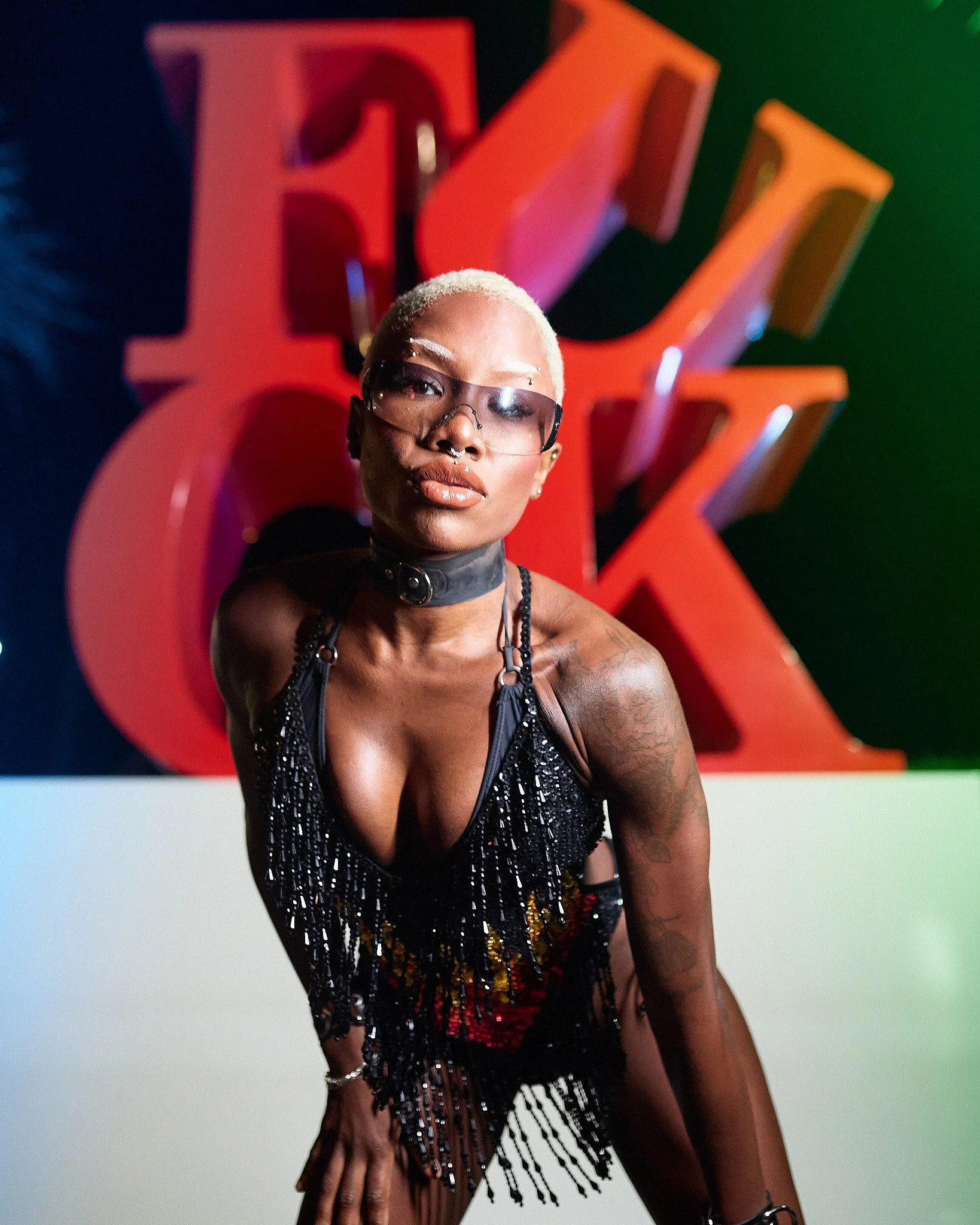 Mujer de cabello rubio platinado con lentes oscuros posando frente a un fondo con letras rojas y amarillas, vistiendo ropa llamativa y de estilo audaz.