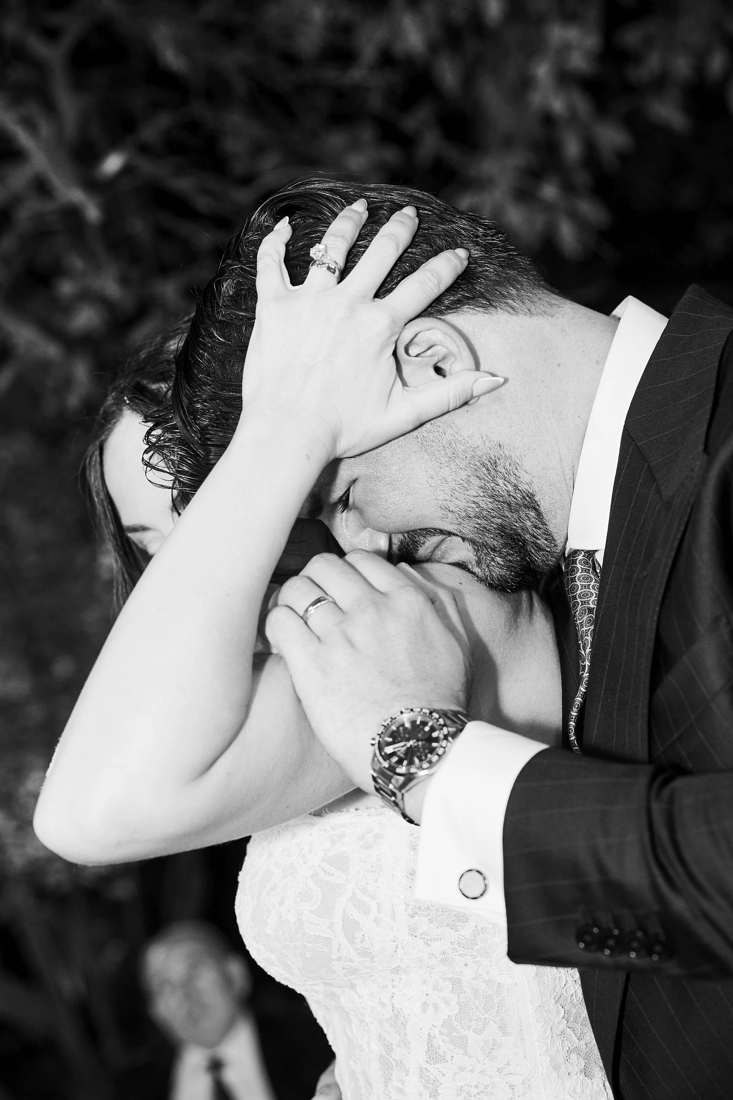 Una pareja de boda, la mujer lleva un vestido de encaje y el hombre un traje formal, están en una pose romántica con las manos entrelazadas y tocándose la cabeza.