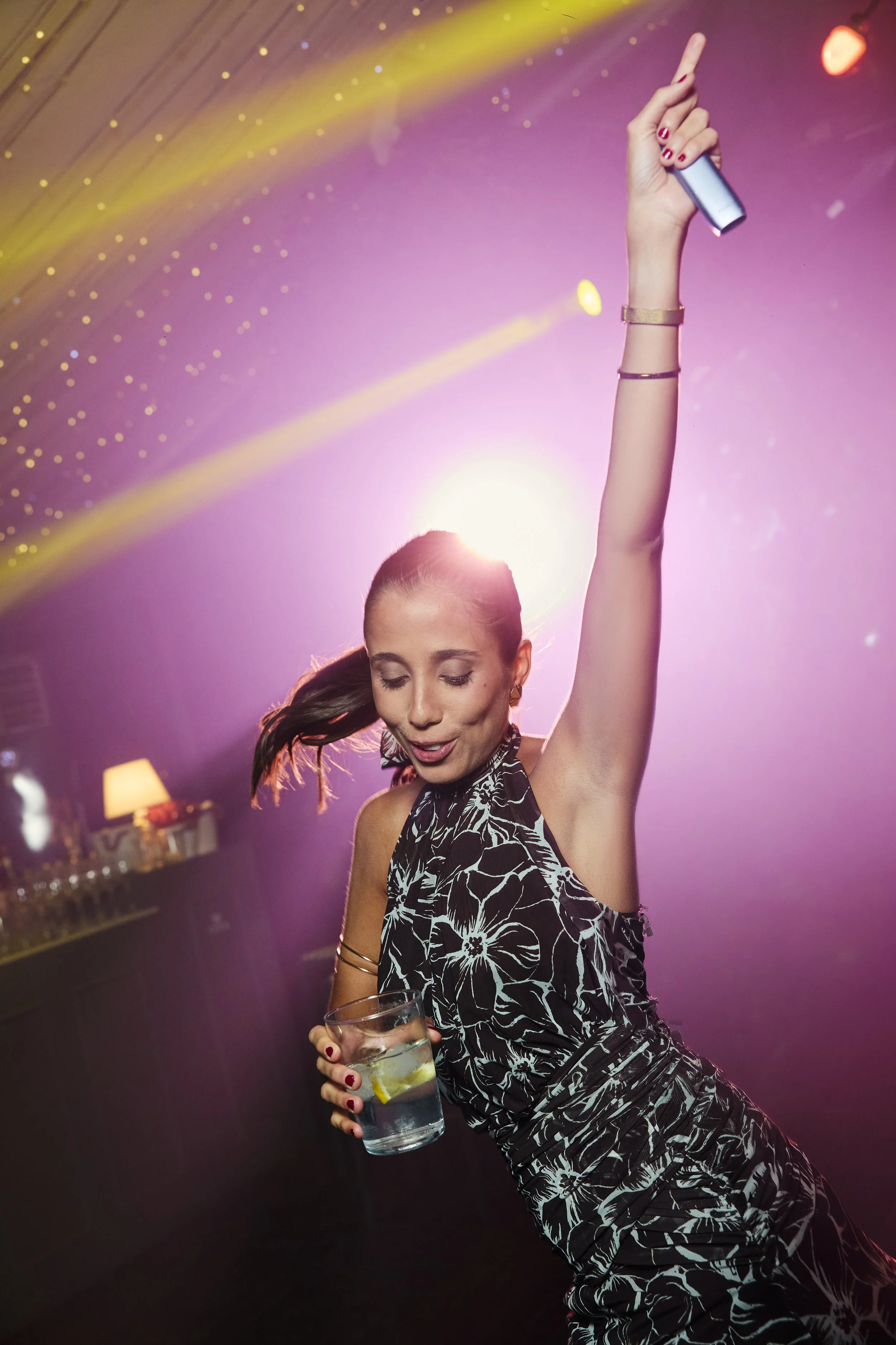 Mujer bailando en un ambiente con luces de colores, sosteniendo un vaso con bebida y decorada con accesorios en la muñeca.
