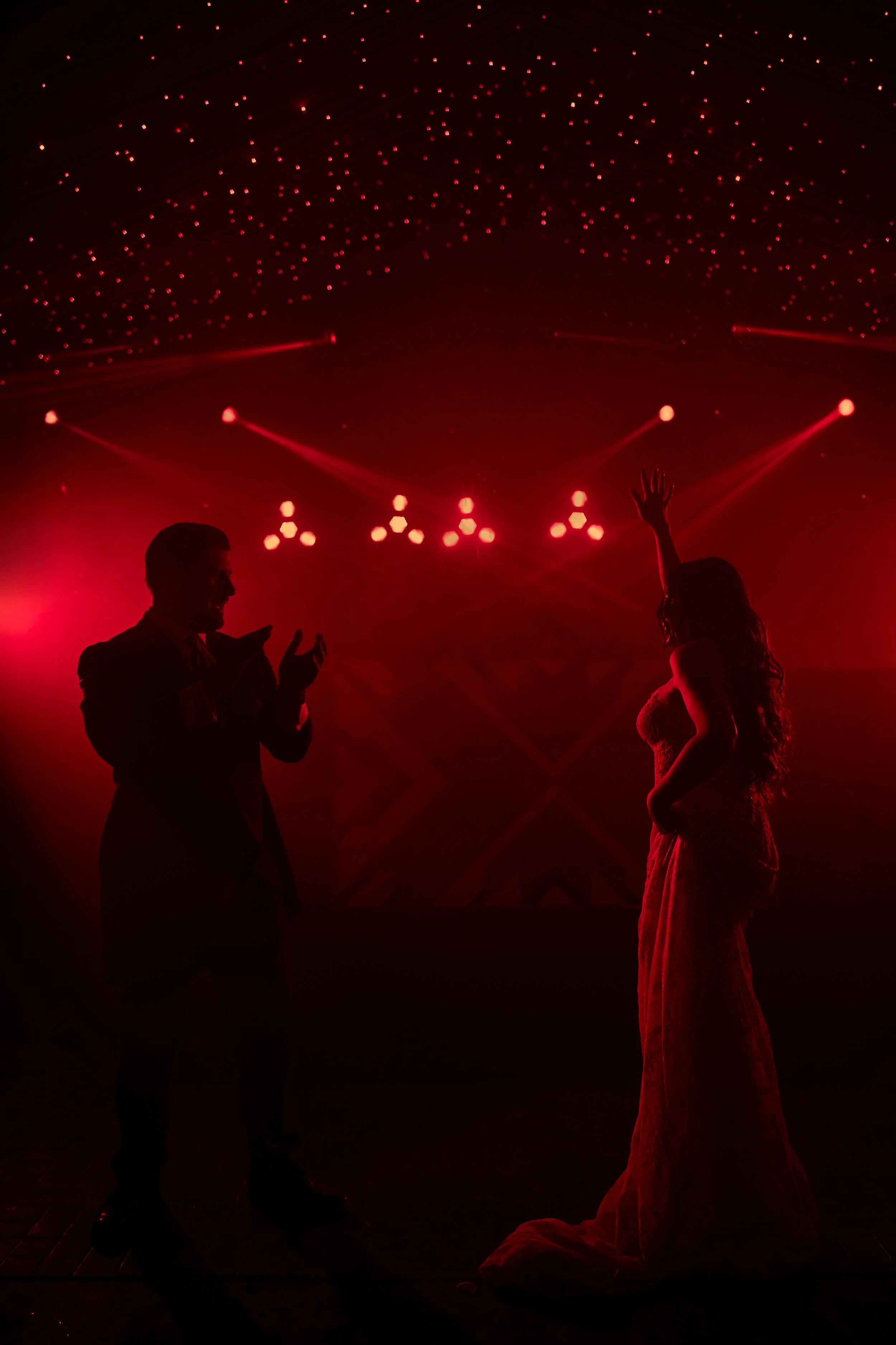 Pareja bailando en una sala con iluminación roja y techos con muchas luces pequeñas, siluetas de un hombre con traje y una mujer con vestido largo.