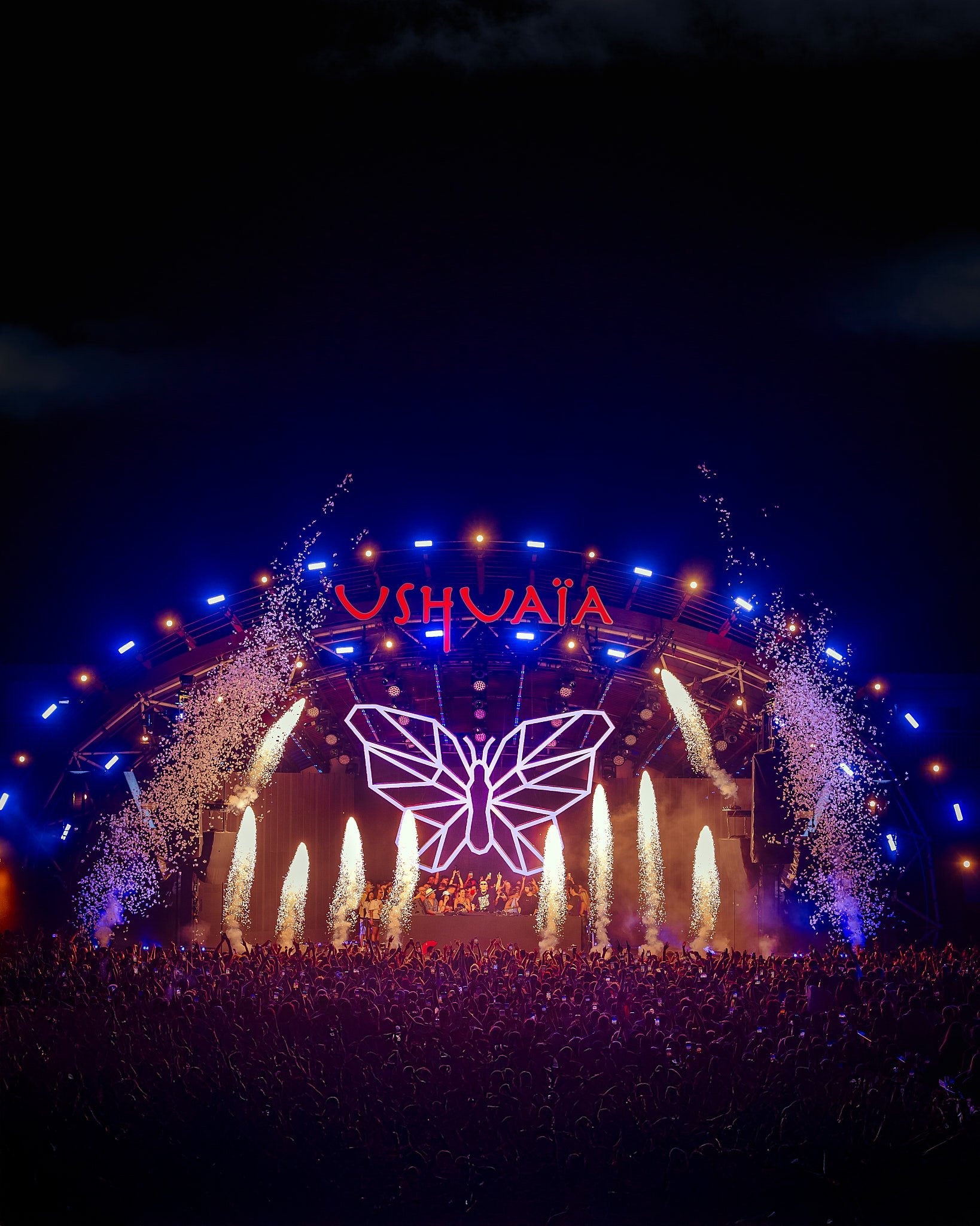 Michael bibi Escenario de concierto con un gran público, decoración de mariposa luminosa en el fondo, fuegos artificiales y la palabra 'USHUAIA' en letras rojas en la parte superior.