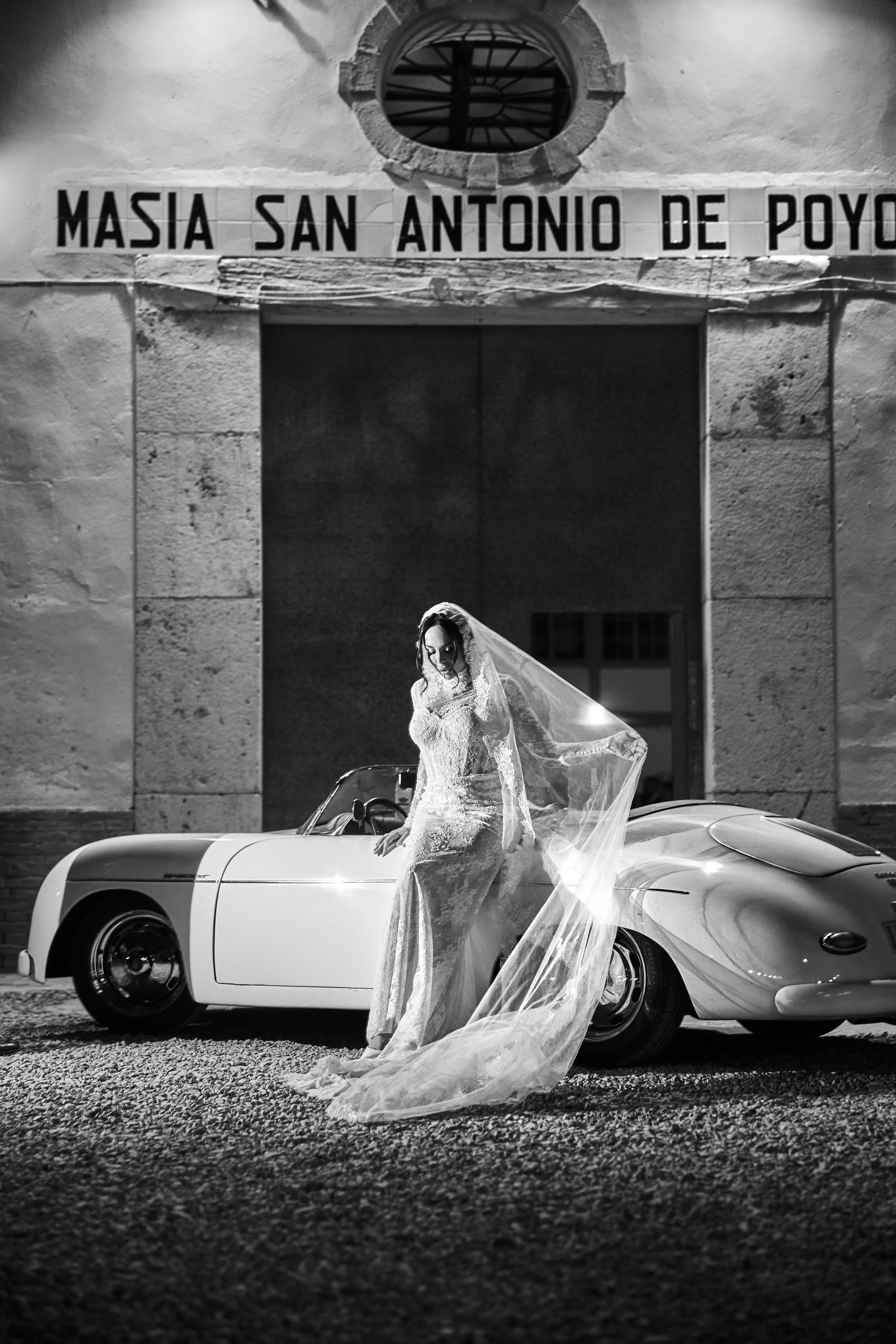 Mujer con vestido de novia y velo posando junto a un coche convertible clásico en una calle de noche, en blanco y negro.
