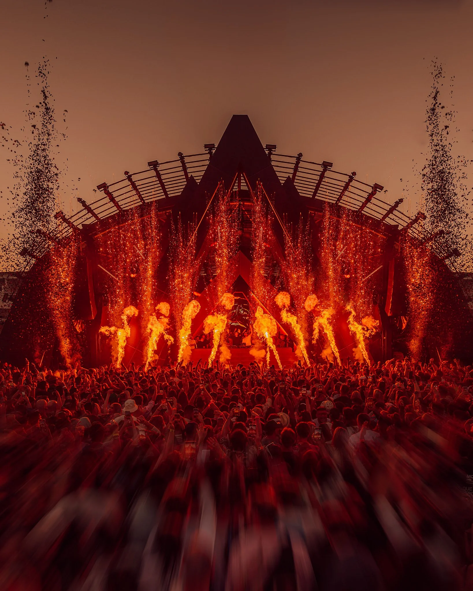Una gran multitud asiste a un concierto al aire libre con llamas y fuegos artificiales en el escenario, en la puesta de sol.