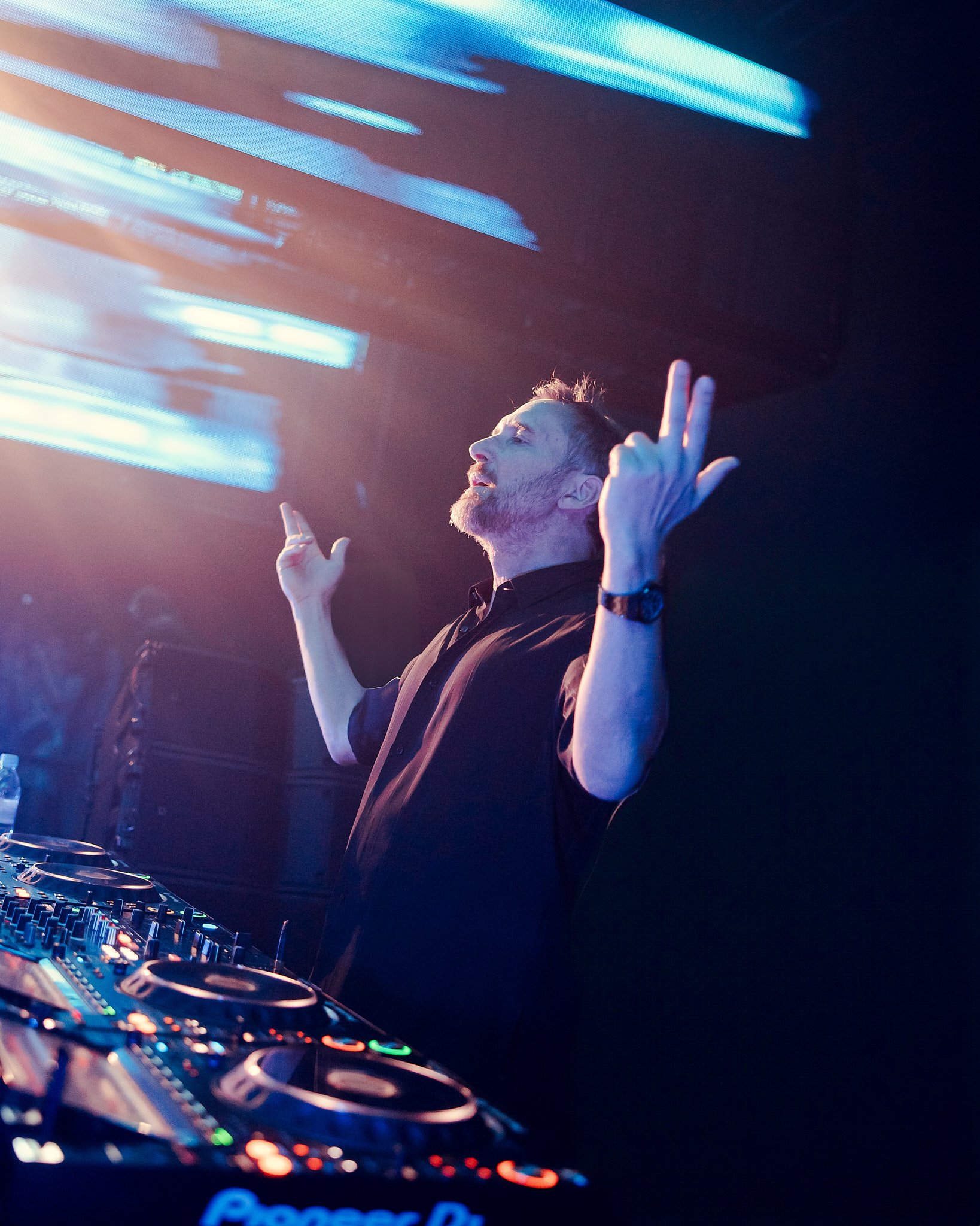 Un DJ en una cabina de disc jockey, mezclando música en un entorno oscuro con luces de neón azules y rosas.