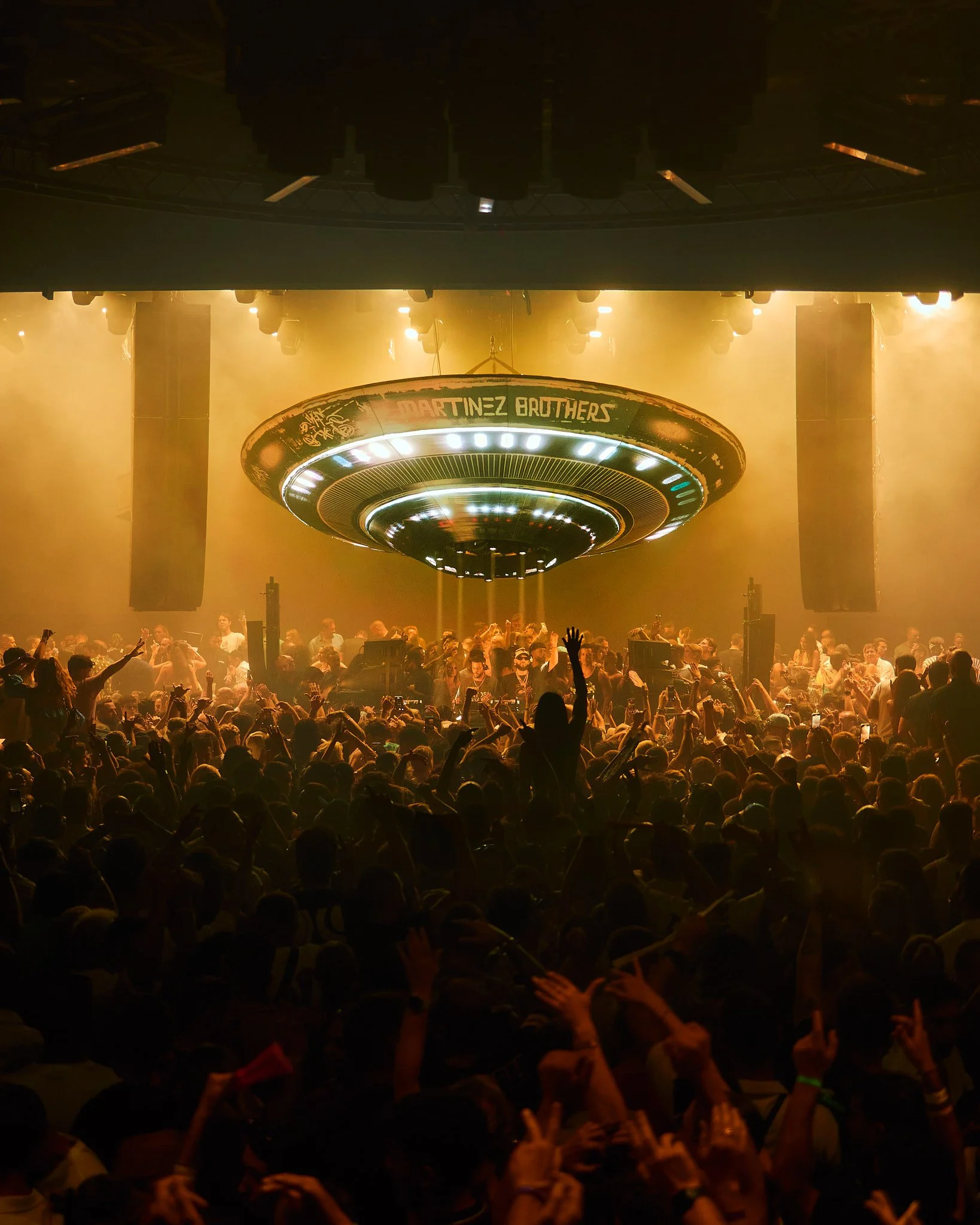 Una multitud en un concierto con luces amarillas y un escenario con un DJ y un cartel que dice 'Martínez Brothers'.