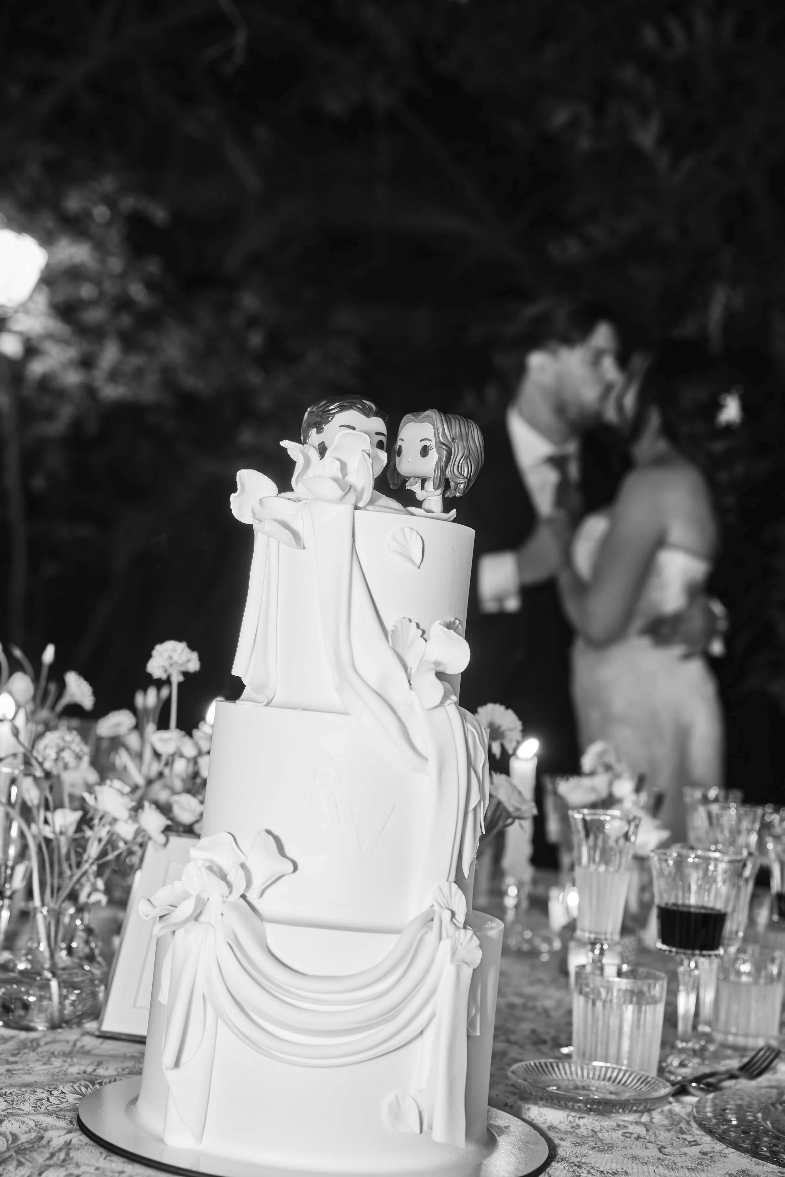 Tarta de boda decorada con figuras de novios en la parte superior, rodeada de flores en una mesa de celebración.