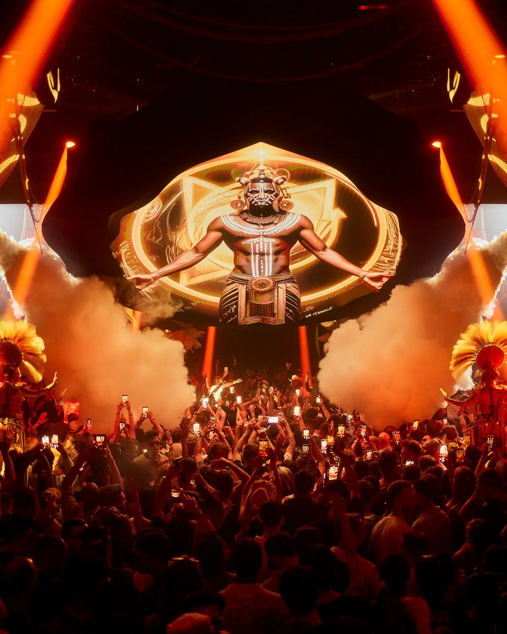 Una multitud en un concierto con efectos de humo y luces, con una pantalla gigante en el fondo que muestra una figura con máscara y accesorios de inspiración africana.
