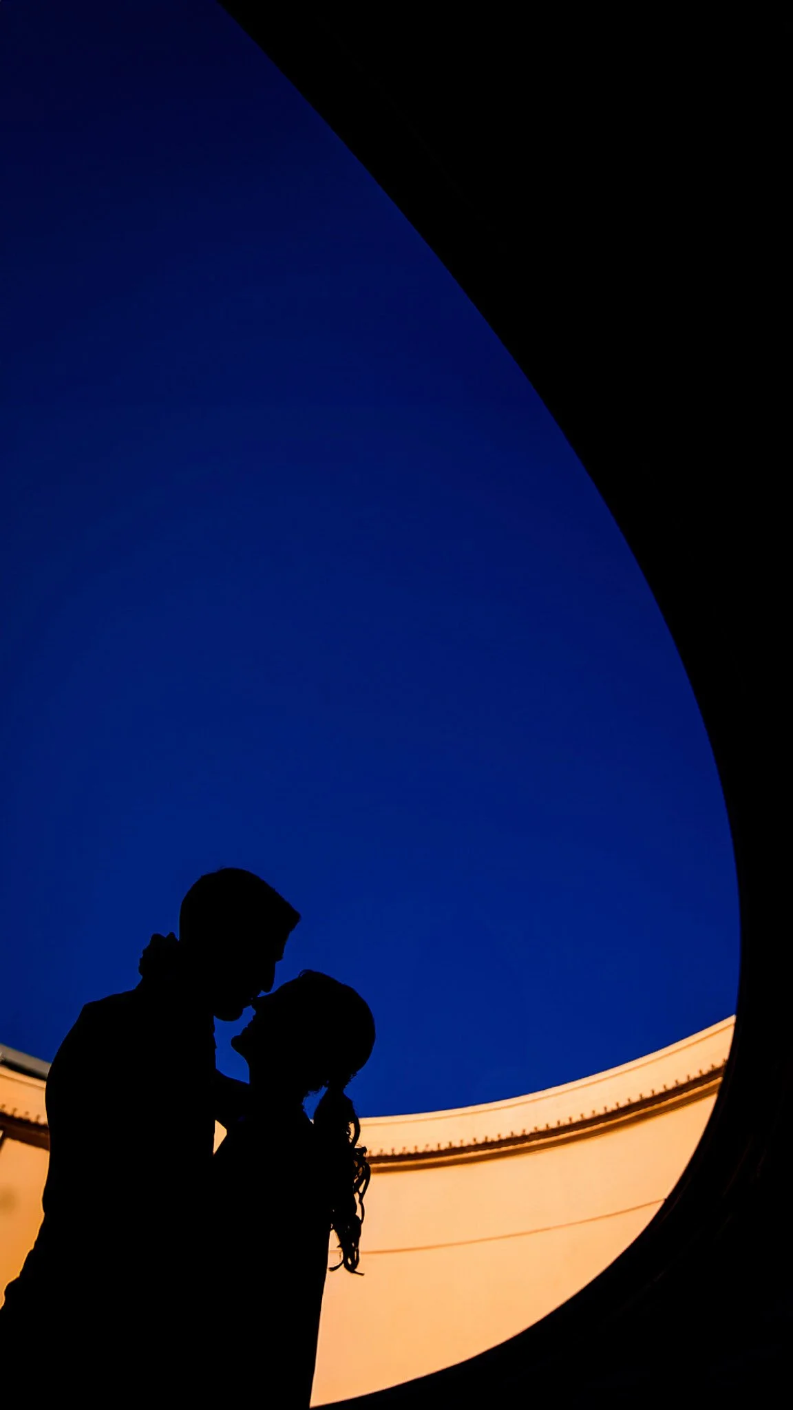 Silhouette de una pareja frente a una estructura circular, con el cielo azul de fondo y una pared anaranjada en la parte inferior.