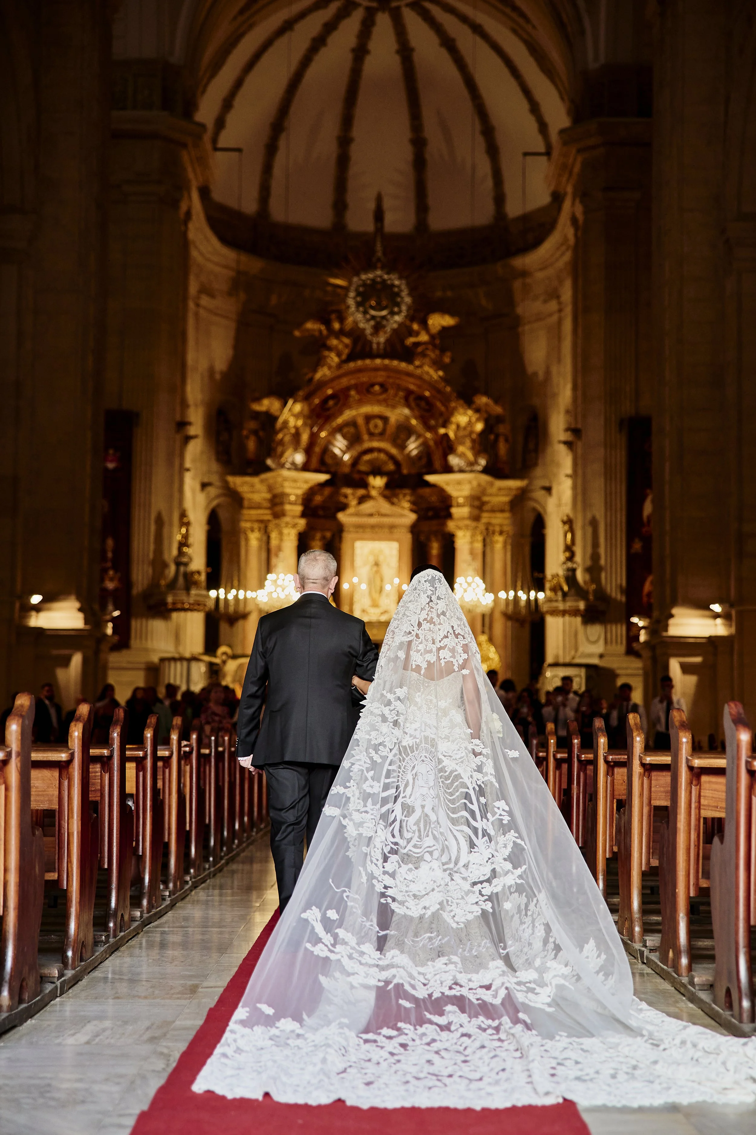 Pareja de novios caminando por el pasillo de una iglesia durante su boda, vista de espaldas, con el altar y candelabros encendidos al fondo.