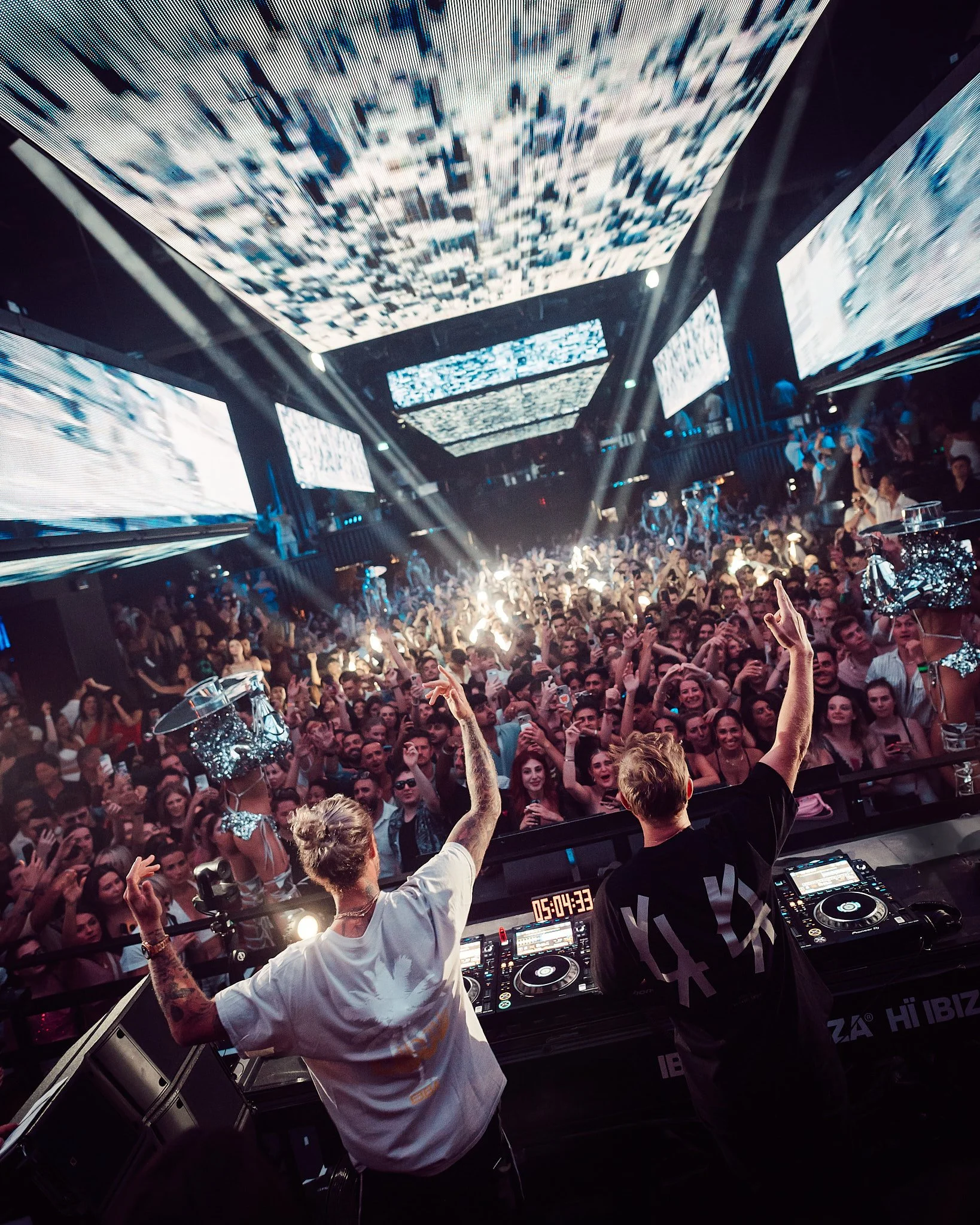 Vista de un club nocturno con una multitud bailando, con dos DJs en el escenario y pantallas enormes con efectos visuales, luces brillantes y ambiente festivo.
