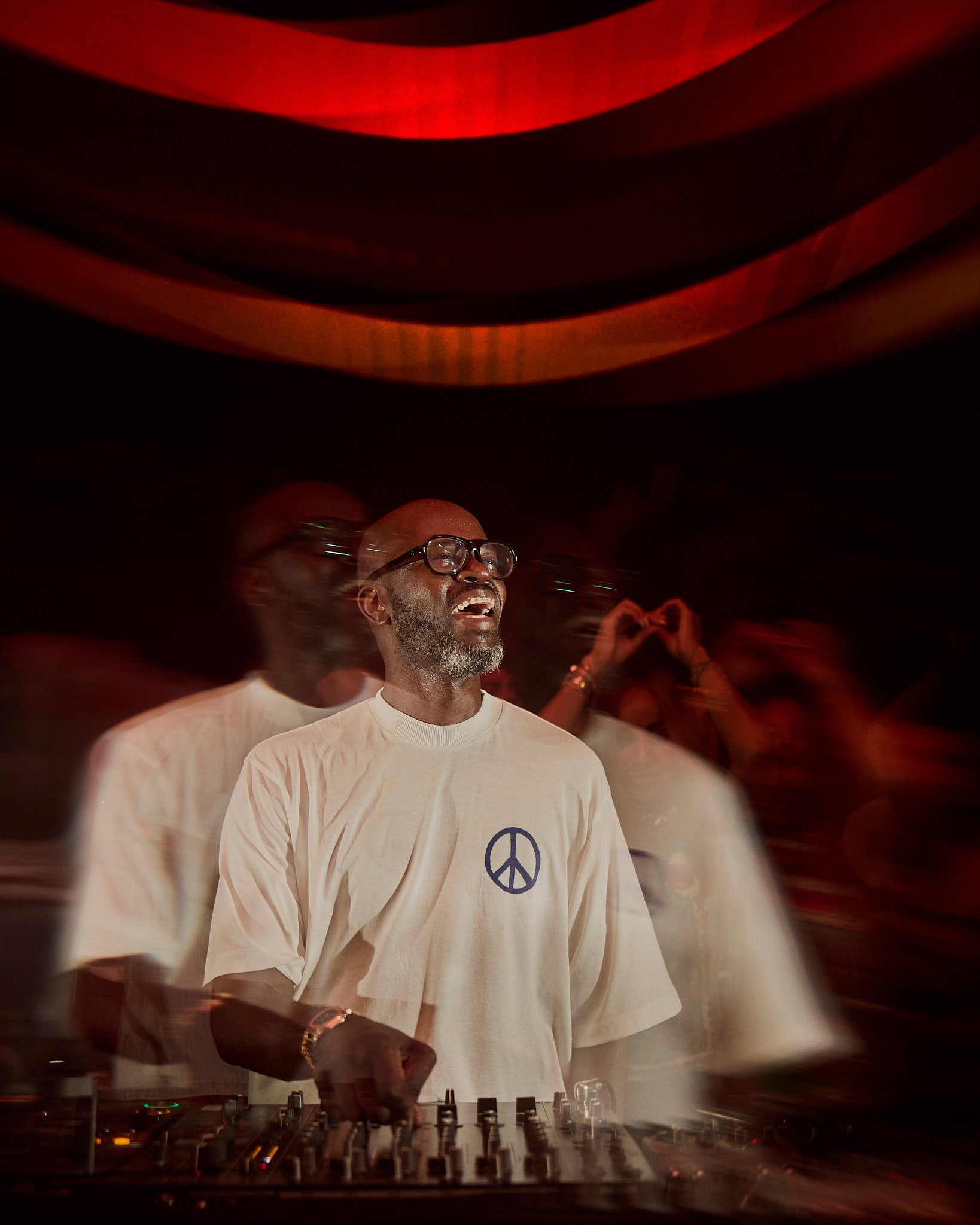 Hombre con gafas y camiseta blanca con símbolo de paz, disfrutando mientras DJ mezcla música en un ambiente con luces rojas.