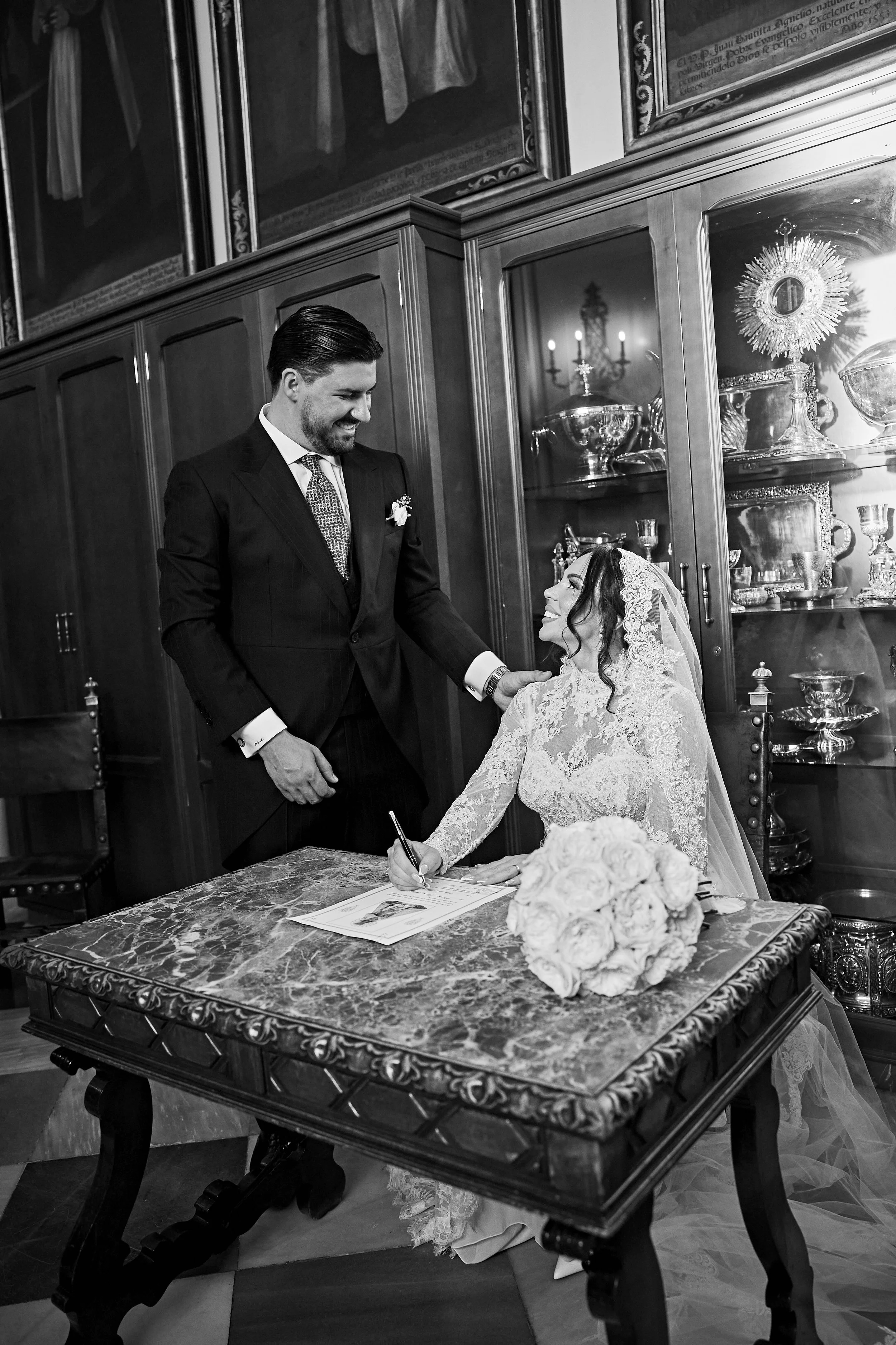 Pareja en una boda, con la mujer firmando un documento y el hombre sonriendo, en una sala con muebles de madera y objetos de plata.