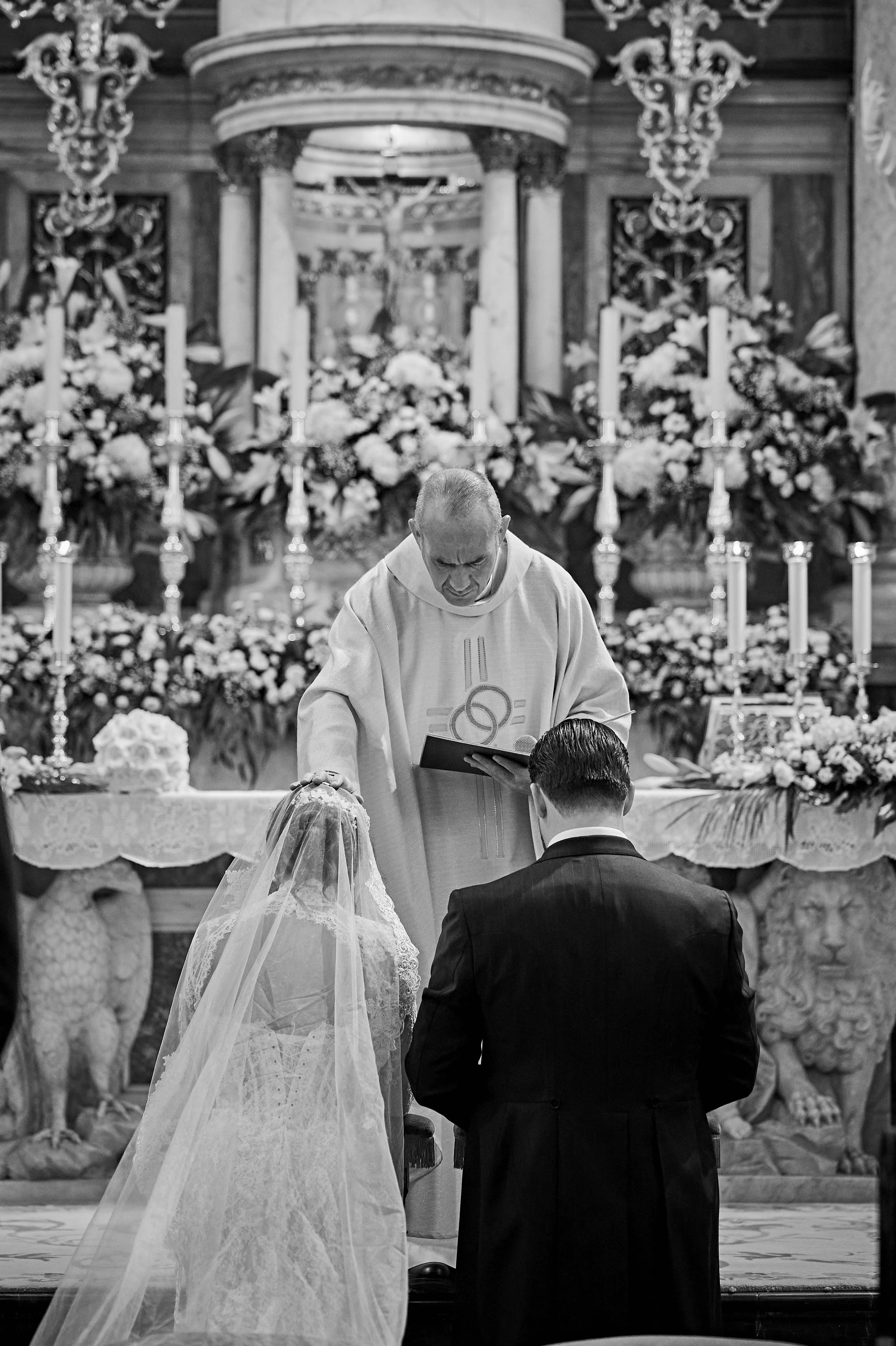 Un sacerdote oficiante en una ceremonia de matrimonio católico, con una pareja en el altar, rodeada de flores y velas, en una iglesia decorada elaboradamente.