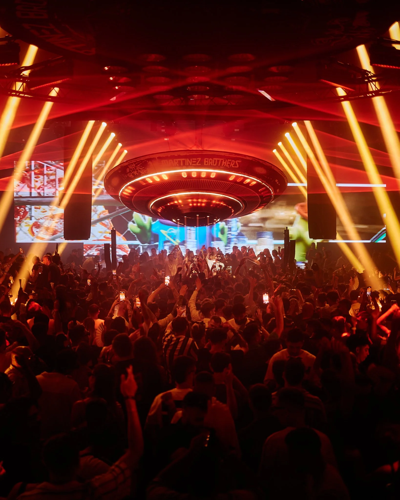 Una multitud de personas en un concierto o evento en un espacio oscuro con muchas luces anaranjadas y pantallas grandes en el fondo que muestran imágenes y videos.