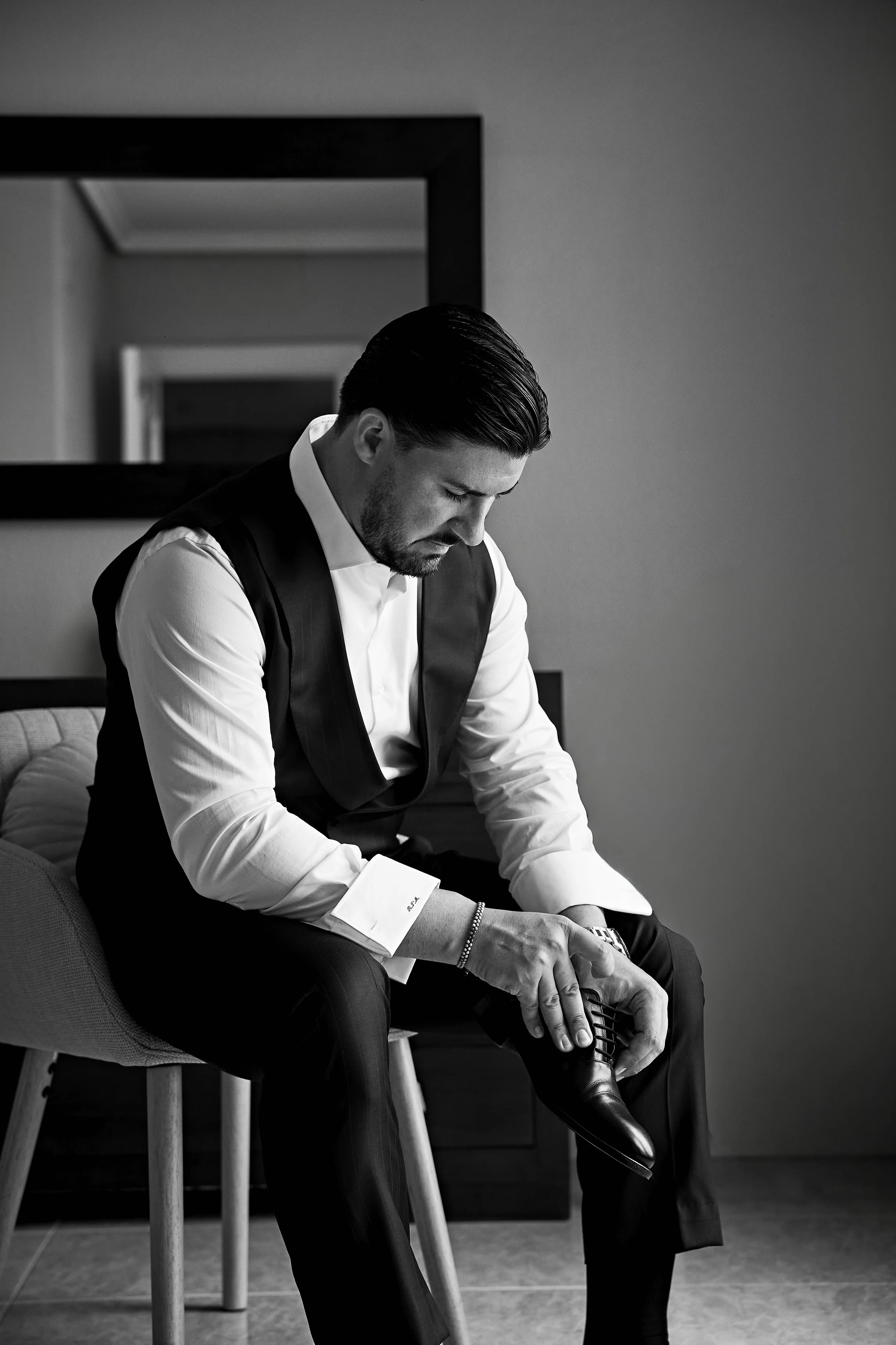 Hombre en ropa formal ajustándose un zapato en una silla en un entorno interior en blanco y negro.