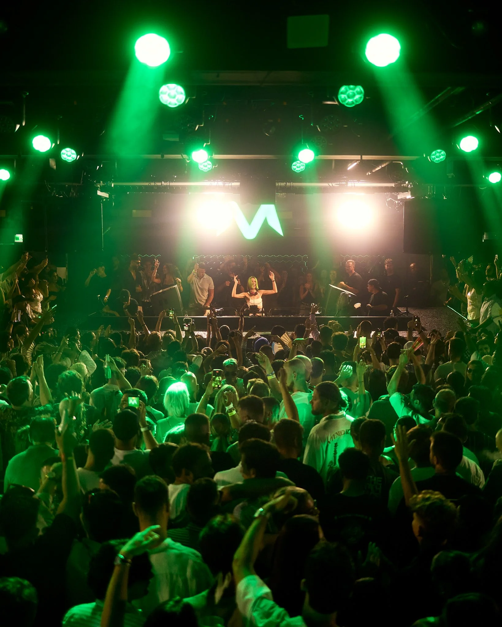 Concierto con público enérgico, luces verdes y escenario con DJ y una pantalla gigante de fondo.