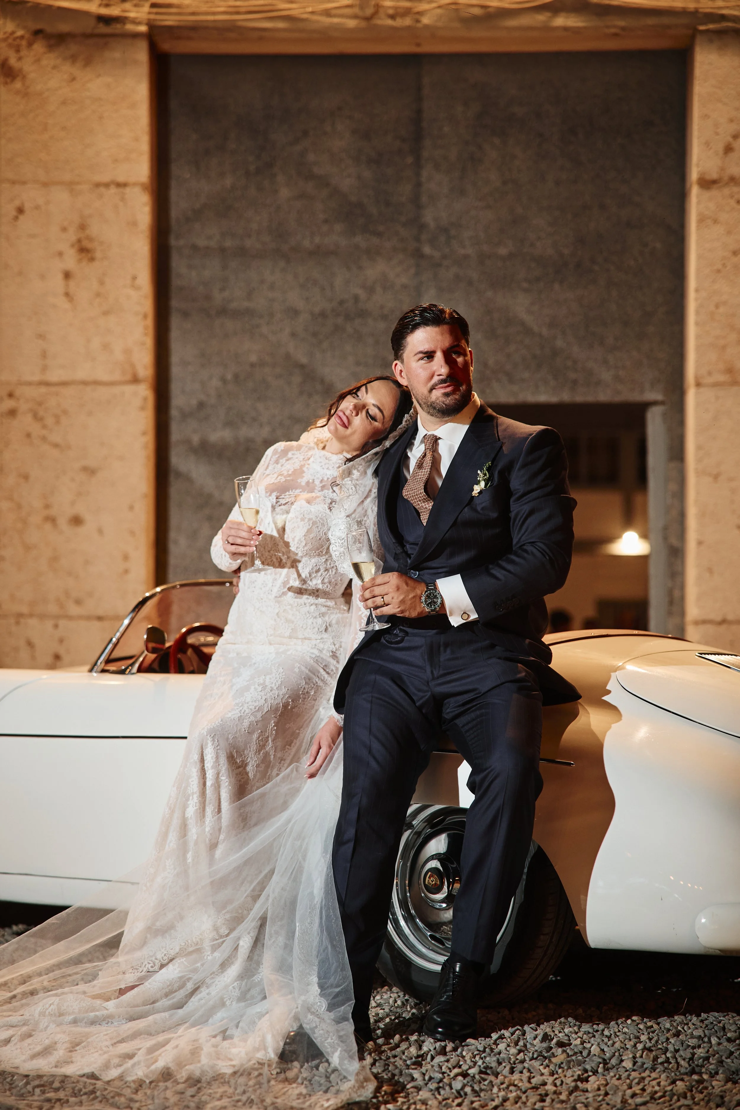 Pareja de recién casados posando junto a un auto clásico blanco. La mujer lleva un vestido de novia de encaje y sostiene una copa de champagne, mientras que el hombre viste un traje oscuro con corbata y reloj. Ambos parecen estar en celebración, en u
