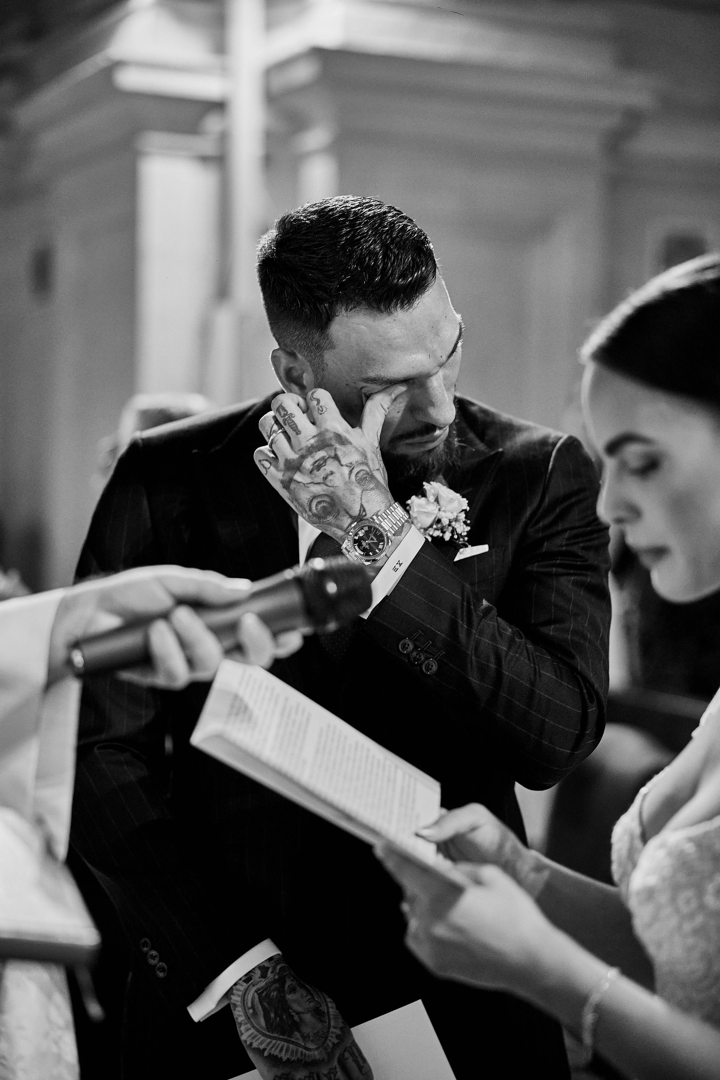 Hombre con tatuajes en la mano, vestido con traje oscuro con flor en el ojal, cubriéndose un ojo con la mano, en una ceremonia o evento formal, acompañado por mujer leyendo algo.