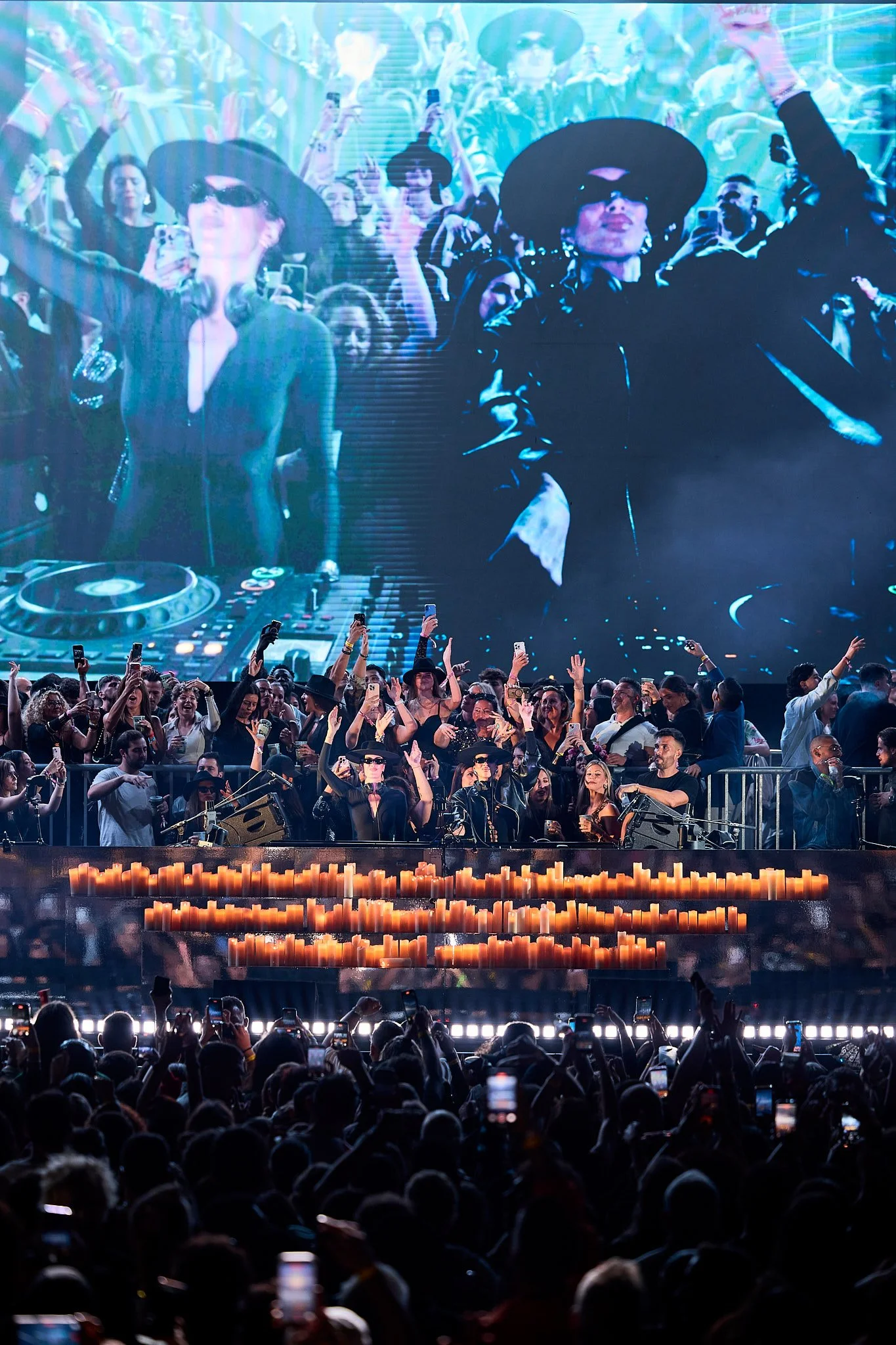 Una multitud en un concierto con un escenario grande, con una pantalla detrás mostrando a dos personas con sombreros y gafas oscuras, y una mesa de DJ. La audiencia está levantando las manos y tomando fotos con sus teléfonos.