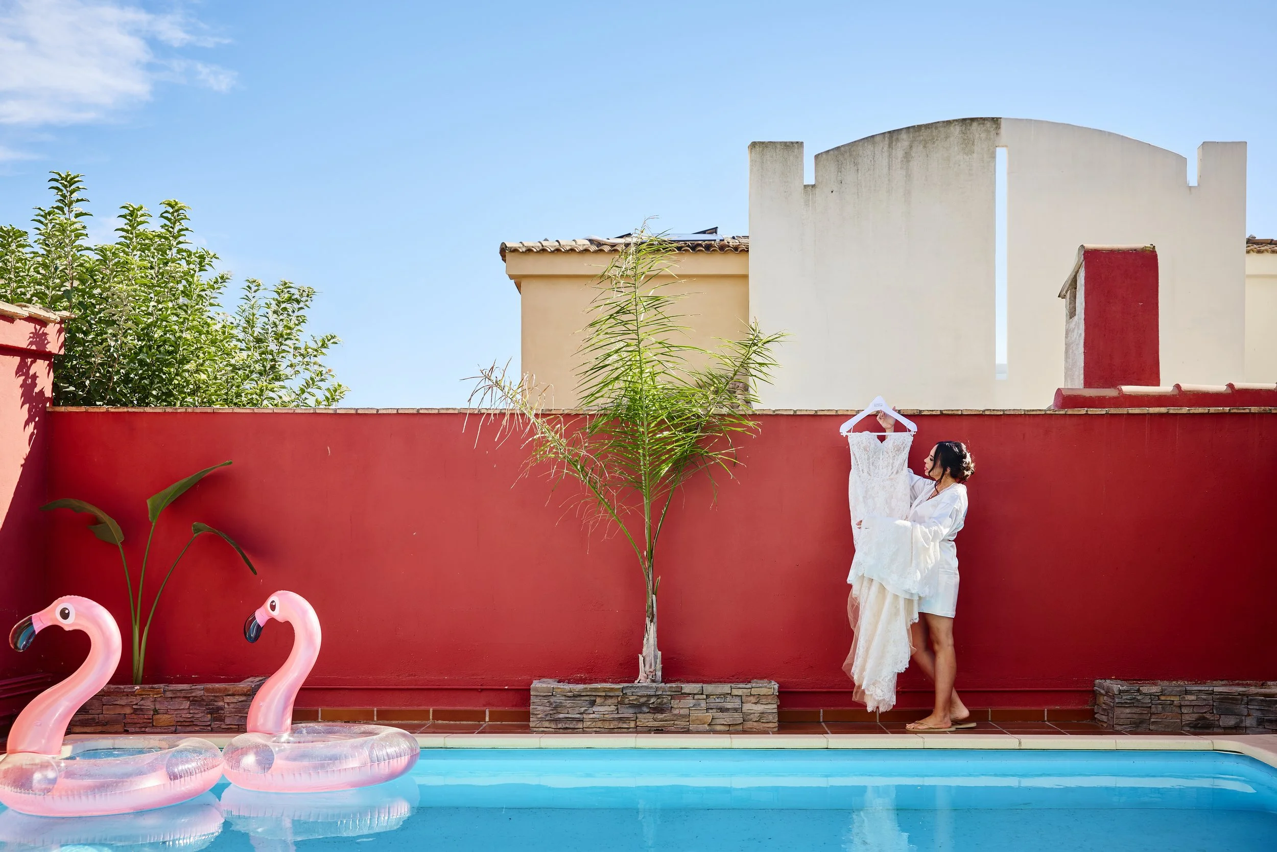 Una mujer con vestido blanco sostiene una prenda de vestir blanca junto a una pared roja cerca de una piscina con flotadores en forma de flamenco y palmeras artificiales, en un fondo de casas con techo de tejas y un cielo despejado.