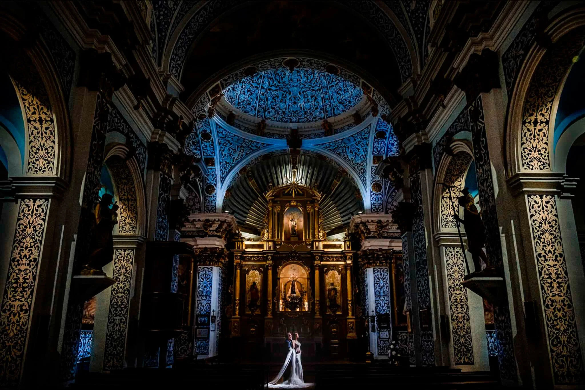 Pareja de novios en el altar de una iglesia, en un espacio decorado con detalles dorados, esculturas y un techo con detalles en azul y blanco.