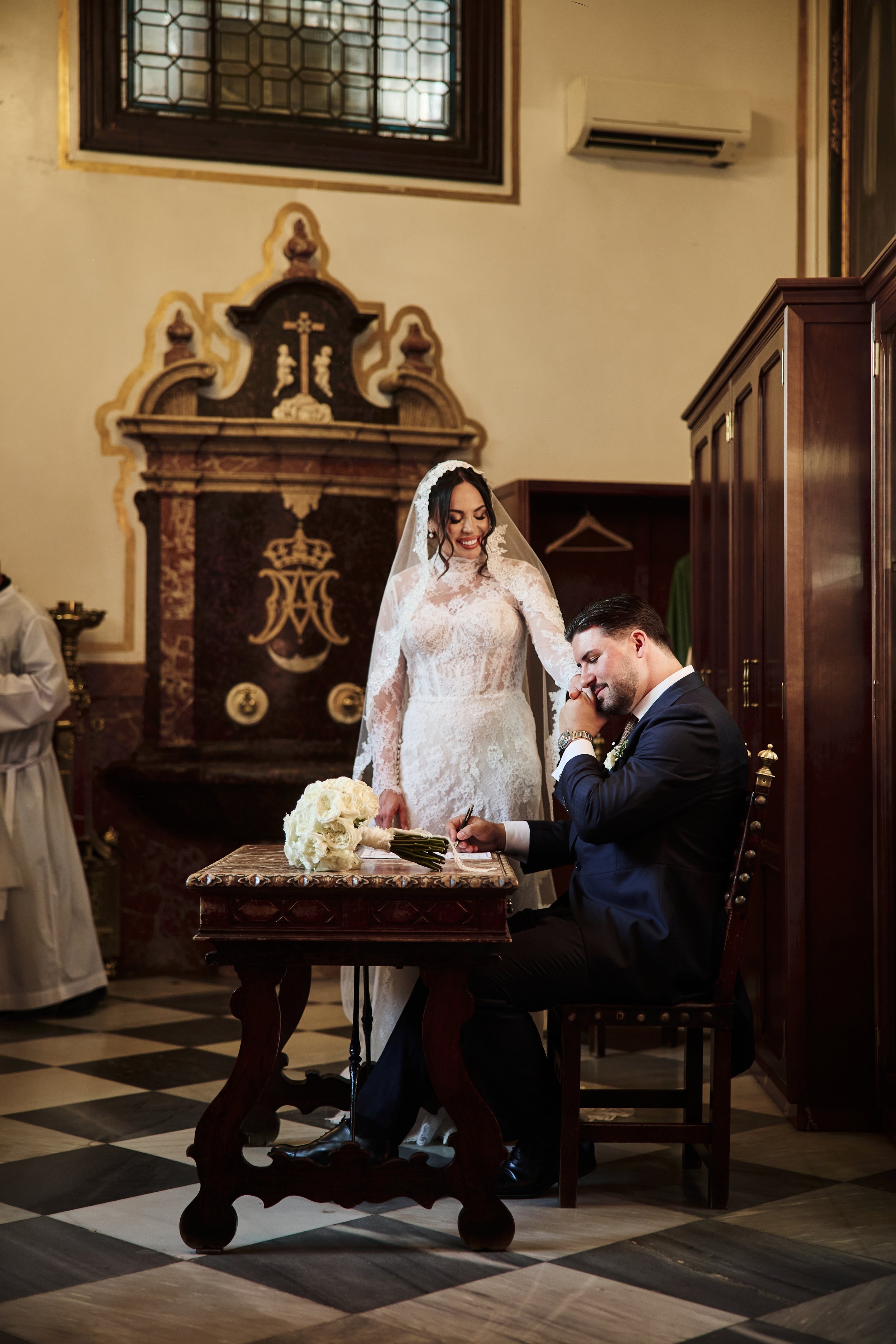 Una mujer con vestido de novia y un hombre con traje gris sentados en una mesa de madera en una ceremonia de boda en un interior elegante, con detalles dorados en el decorado y un órgano en la pared de fondo.