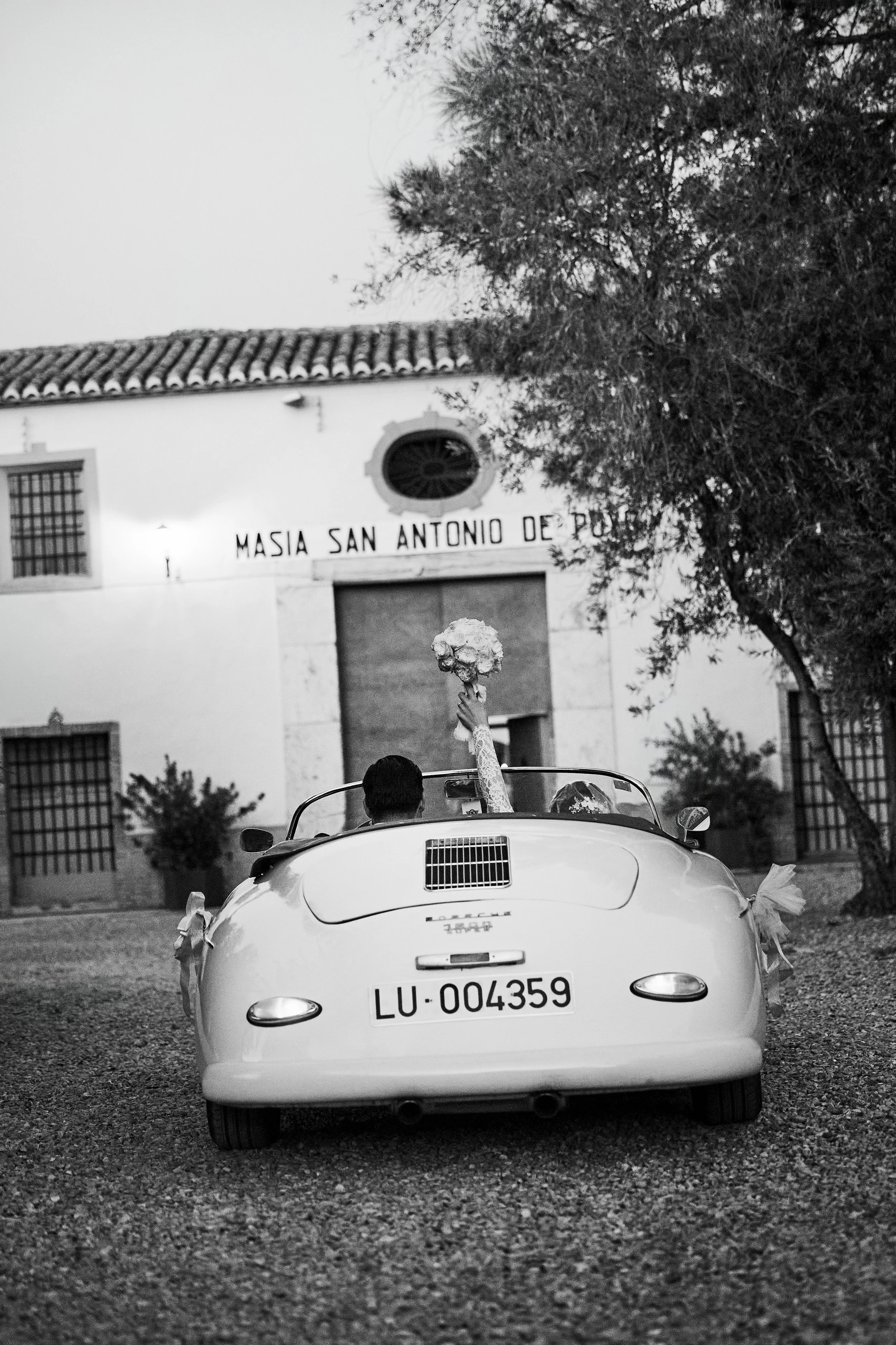 Un coche clásico blanco de dos plazas con matrícula LU-004359, en un entorno de piedra y césped, con un árbol y un edificio antiguo con puerta grande y ventanas con rejas, y un cartel que dice 'MASIA SAN ANTONIO DE PUEBLO'.
