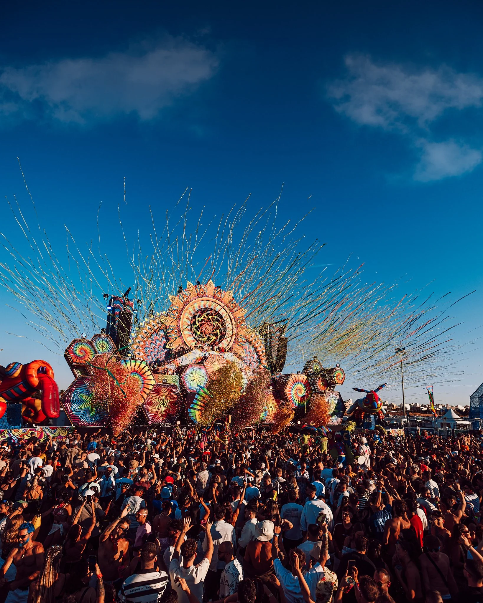 ELROW con un escenario decorado con luces y figuras coloridas, mucha gente disfrutando del evento en un día soleado.