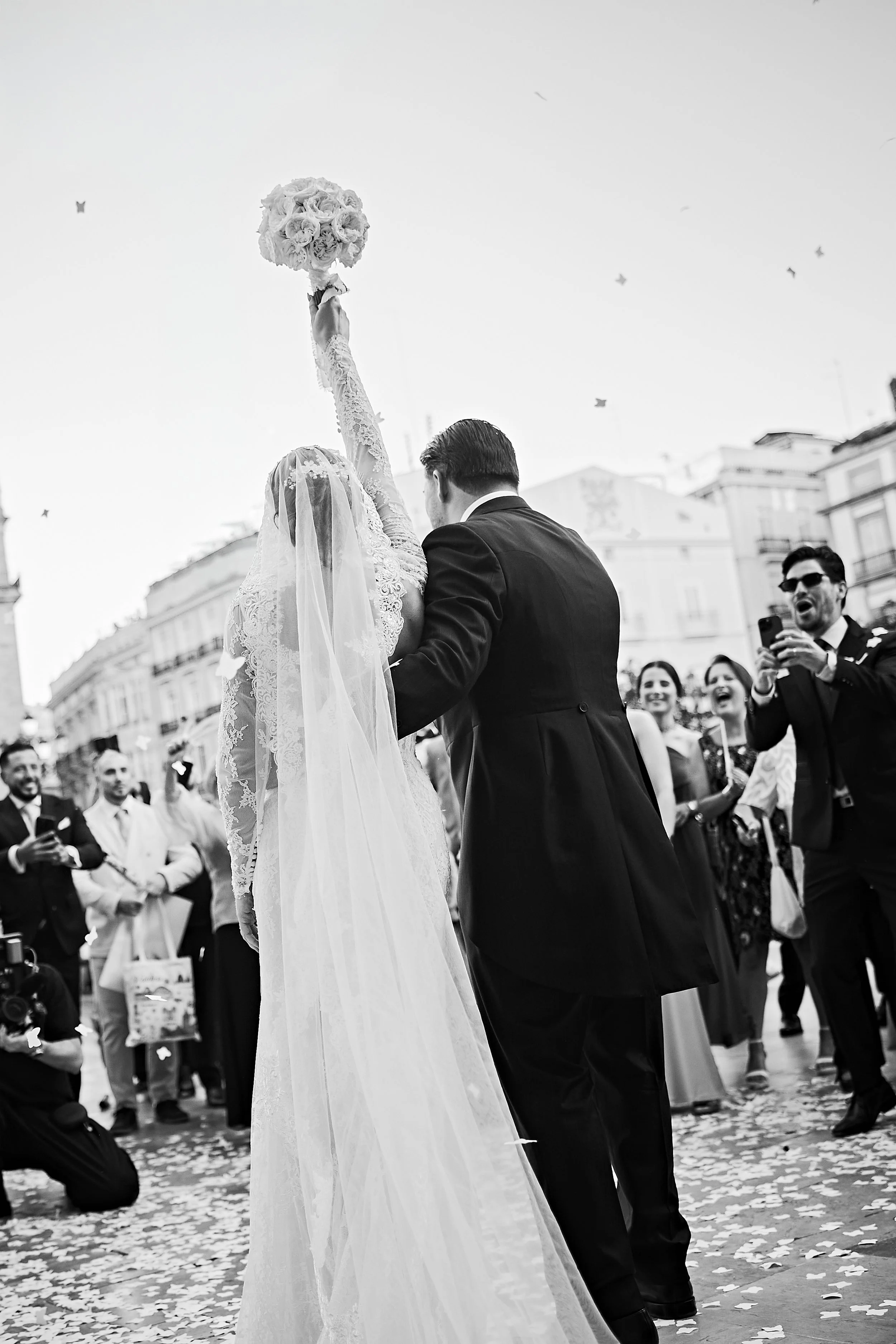 Pareja de novios en una boda, la novia sostiene un ramo de rosas en alto, rodeada de invitados en la calle, con confeti en el suelo y algunas personas capturando el momento con sus teléfonos.