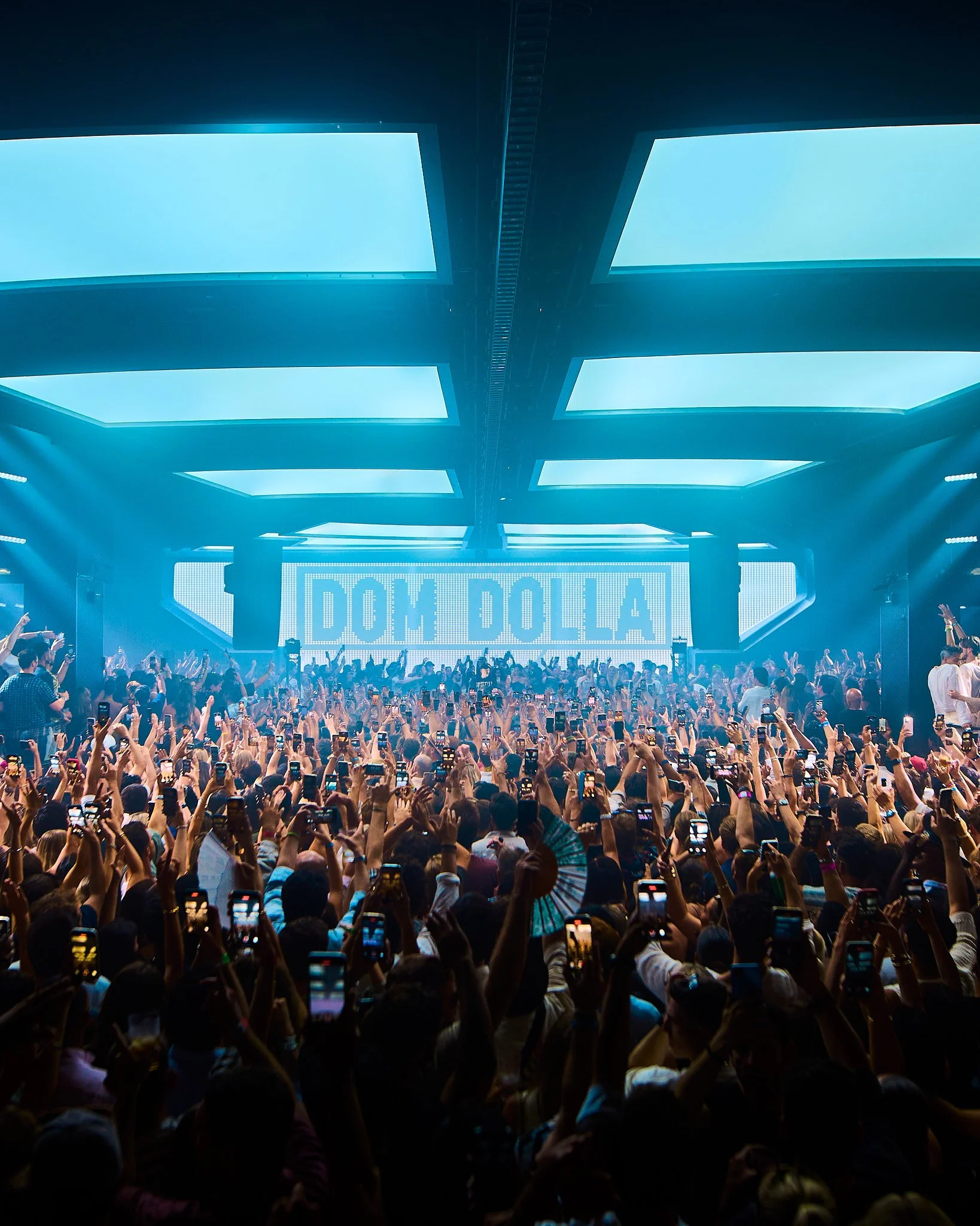 Concierto con gran multitud en el escenario con letras que dicen 'DOM DOLLA' y espectadores tomando fotos con teléfonos.