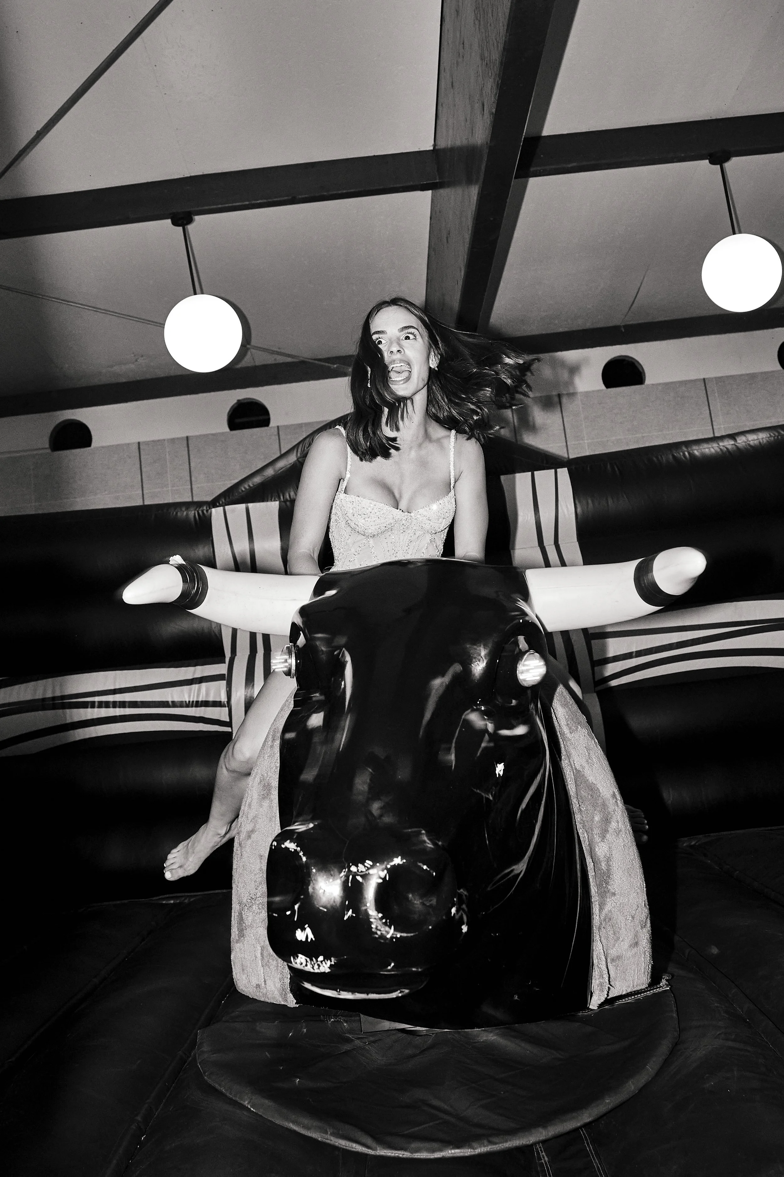 Una mujer con cabello suelto y expresión de diversión en un juego mecánico en forma de toro con cabeza de toro brillante y cuernos blancos, en un ambiente con luces redondas en el techo.