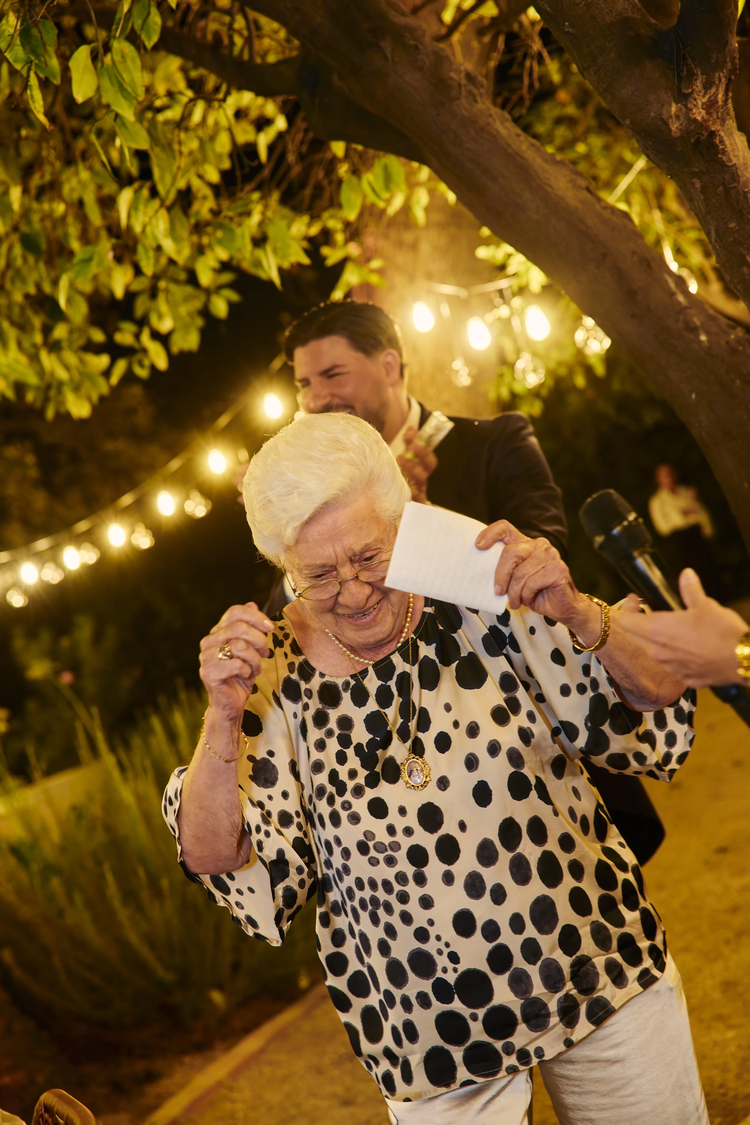 Una mujer mayor con blusa de lunares negros y blancos riendo y disfrutando en una celebración al aire libre con luces amarillas y un hombre joven en el fondo.