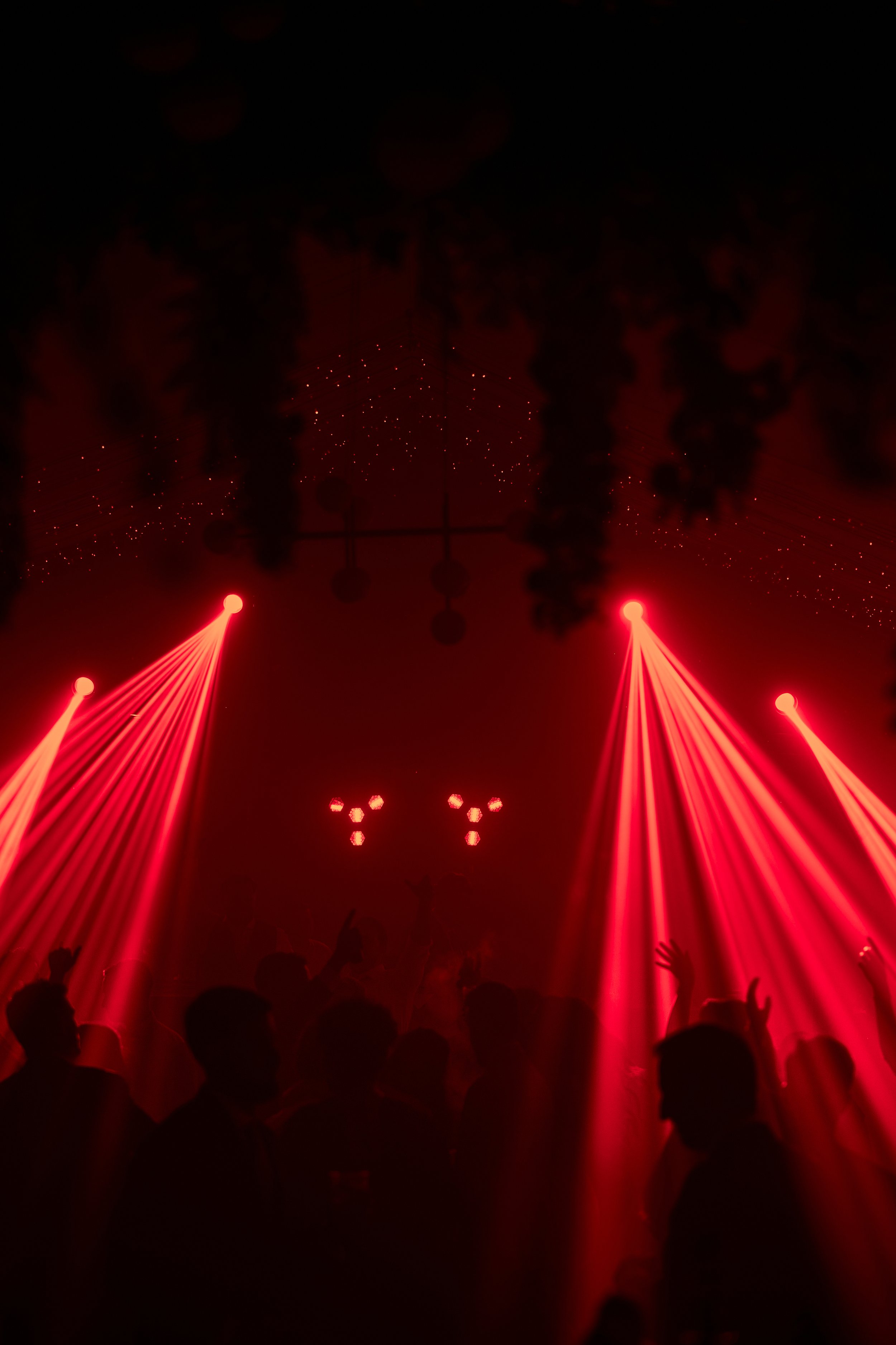 Se observan luces rojas en un ambiente oscuro, con personas bailando y levantando las manos en un concierto o fiesta con un espectáculo de luces.