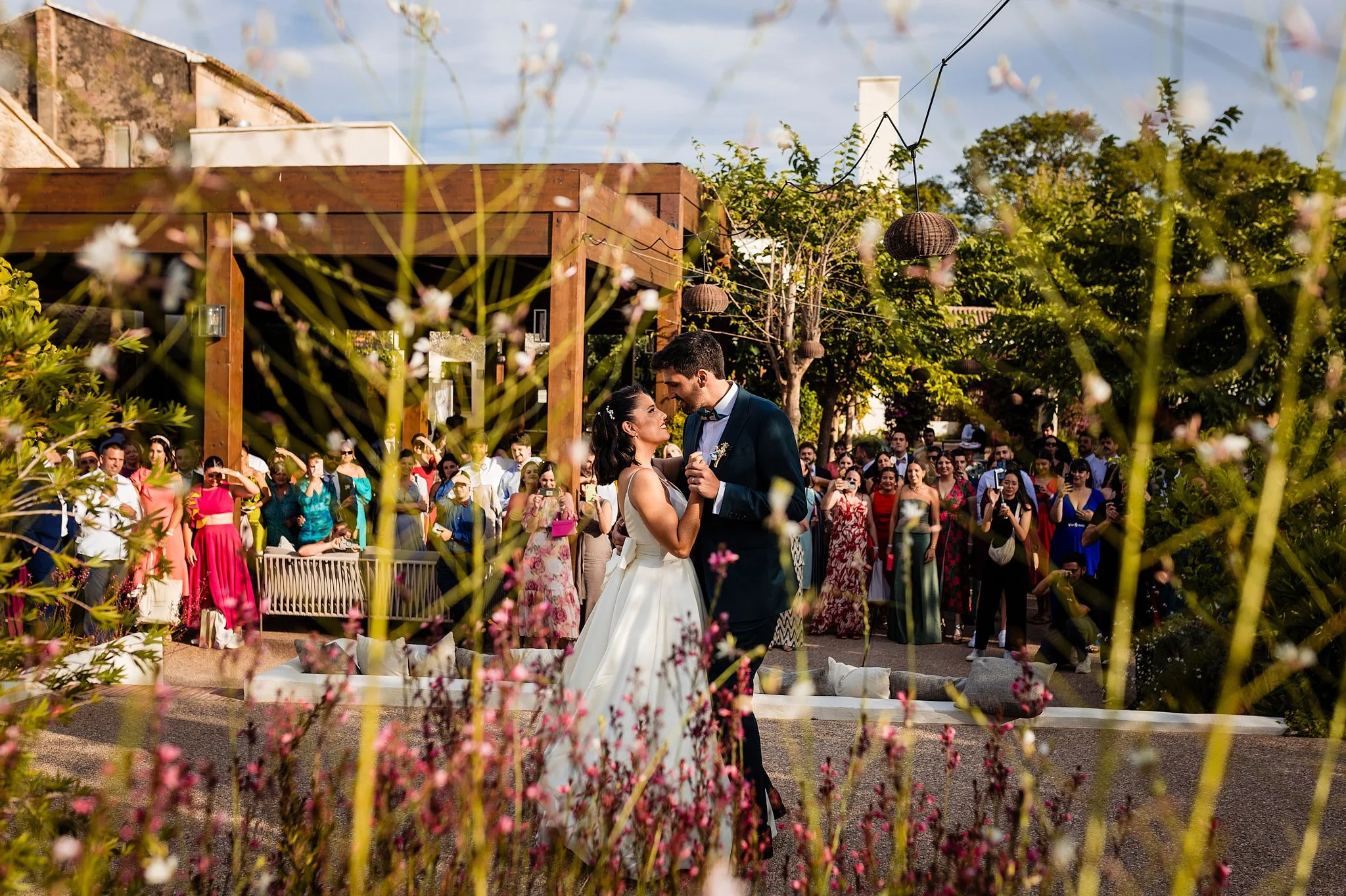 Pareja de recién casados bailando en una recepción al aire libre, rodeados de invitados y vegetación, en un ambiente de celebración y alegría.