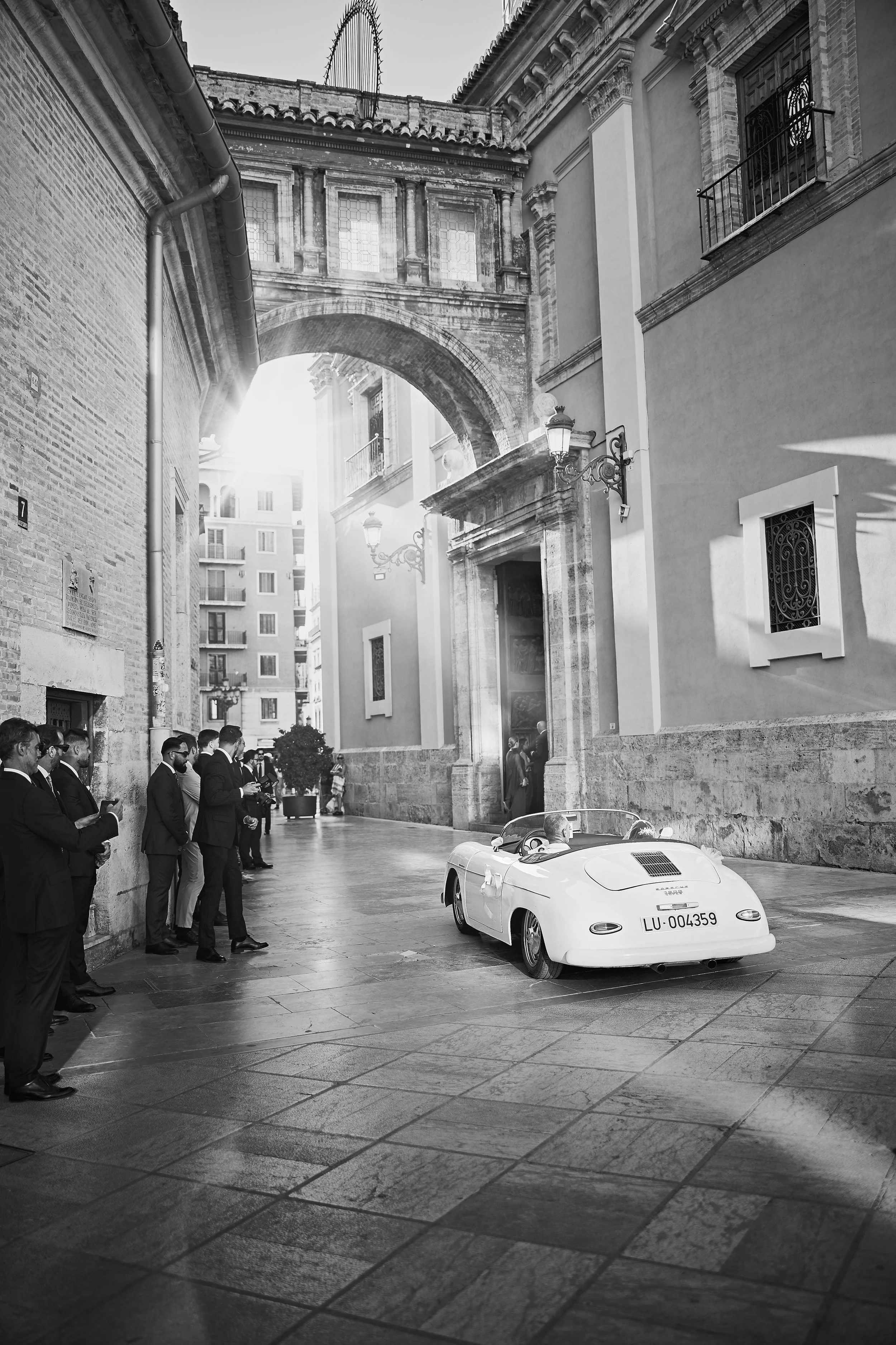 Escena en una calle urbana con varias personas vestidas con traje, un automóvil deportivo blanco y arquitectura antigua en el fondo.