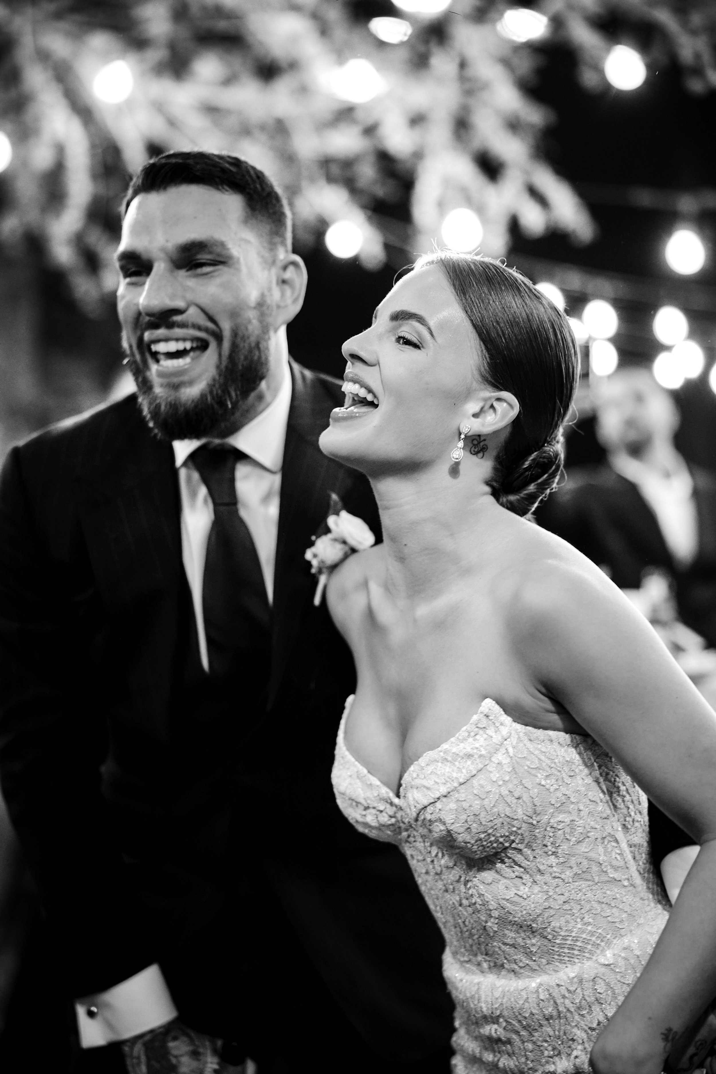 Pareja de novios riendo en una boda, la mujer lleva vestido de encaje y aretes largos, el hombre viste traje y corbata, ambiente con luces decorativas.