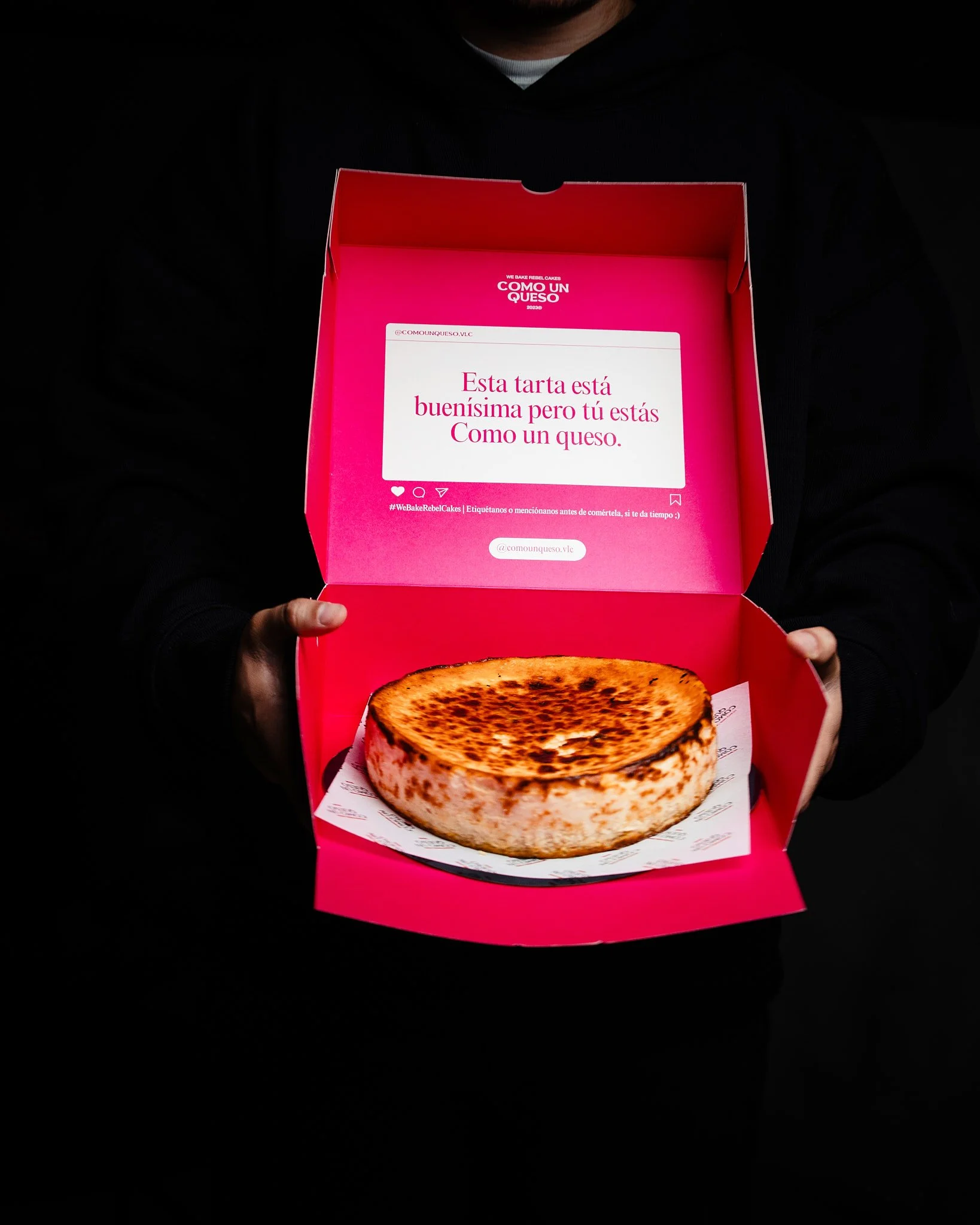 Una caja de pastel de queso con el interior rosa que contiene un pastel de queso tostado. En la tapa de la caja, hay un mensaje que dice que la tarta está buenísima pero tú eres como un queso.