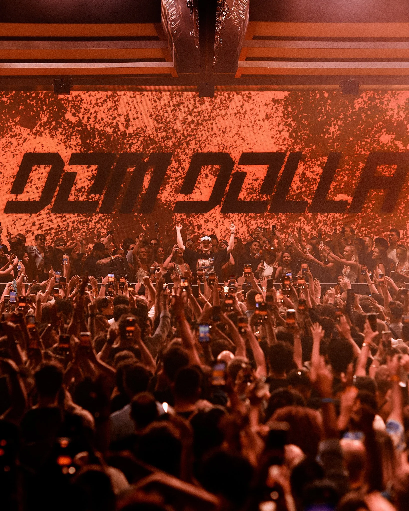 Escenario con logo de DJ en fondo de luces rojas, con multitud de personas disfrutando, muchas con teléfonos en mano.