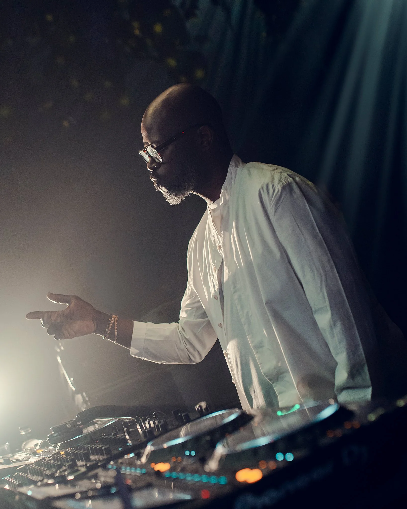 Un DJ con barba y gafas de sol, vestido con una camisa blanca, mezclando música en una cabina con luces ambientales.