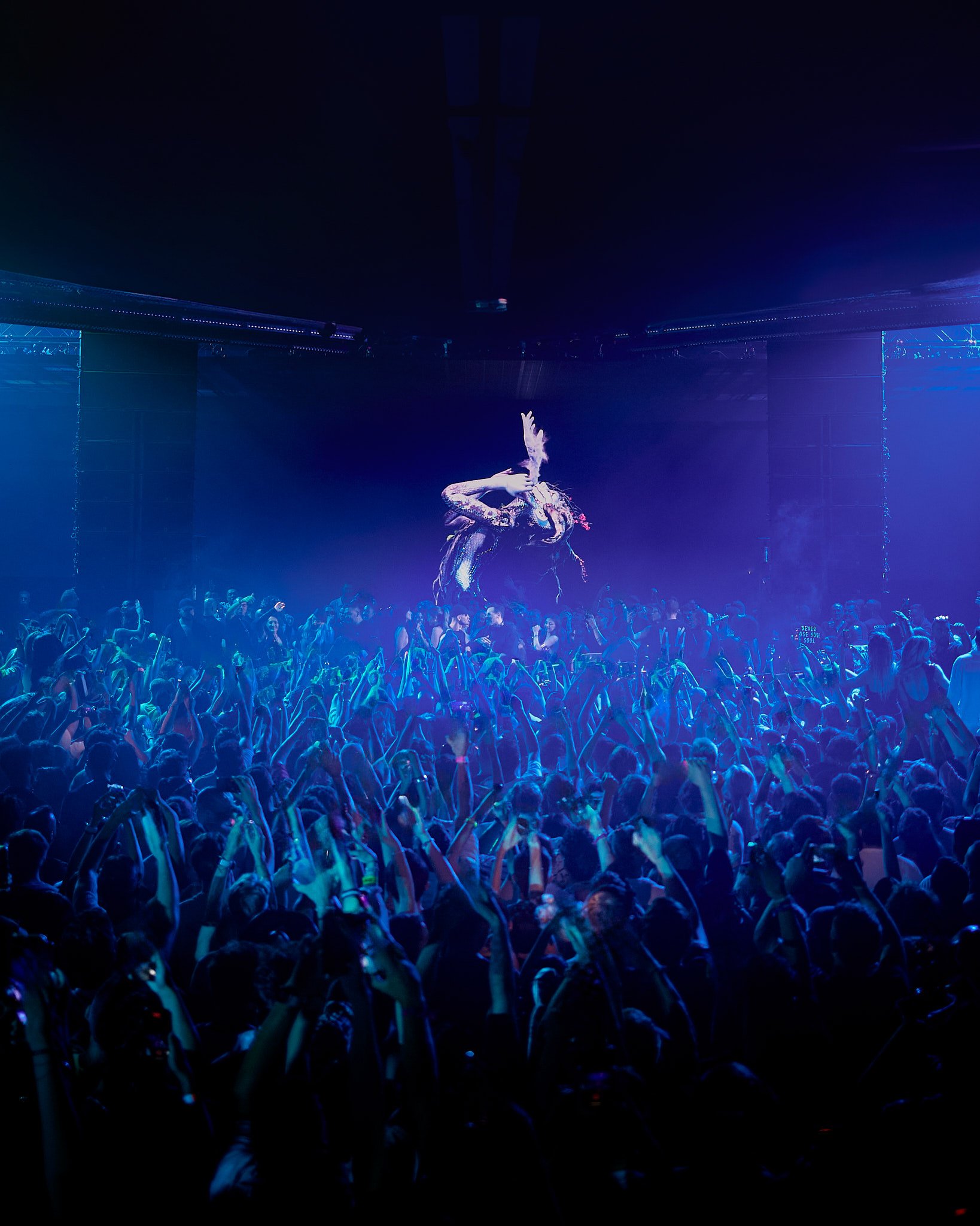 Concierto con un escenario iluminado en tonos azules y una multitud de personas bailando y levantando las manos, en el centro hay una artista cantando con un fondo oscuro.